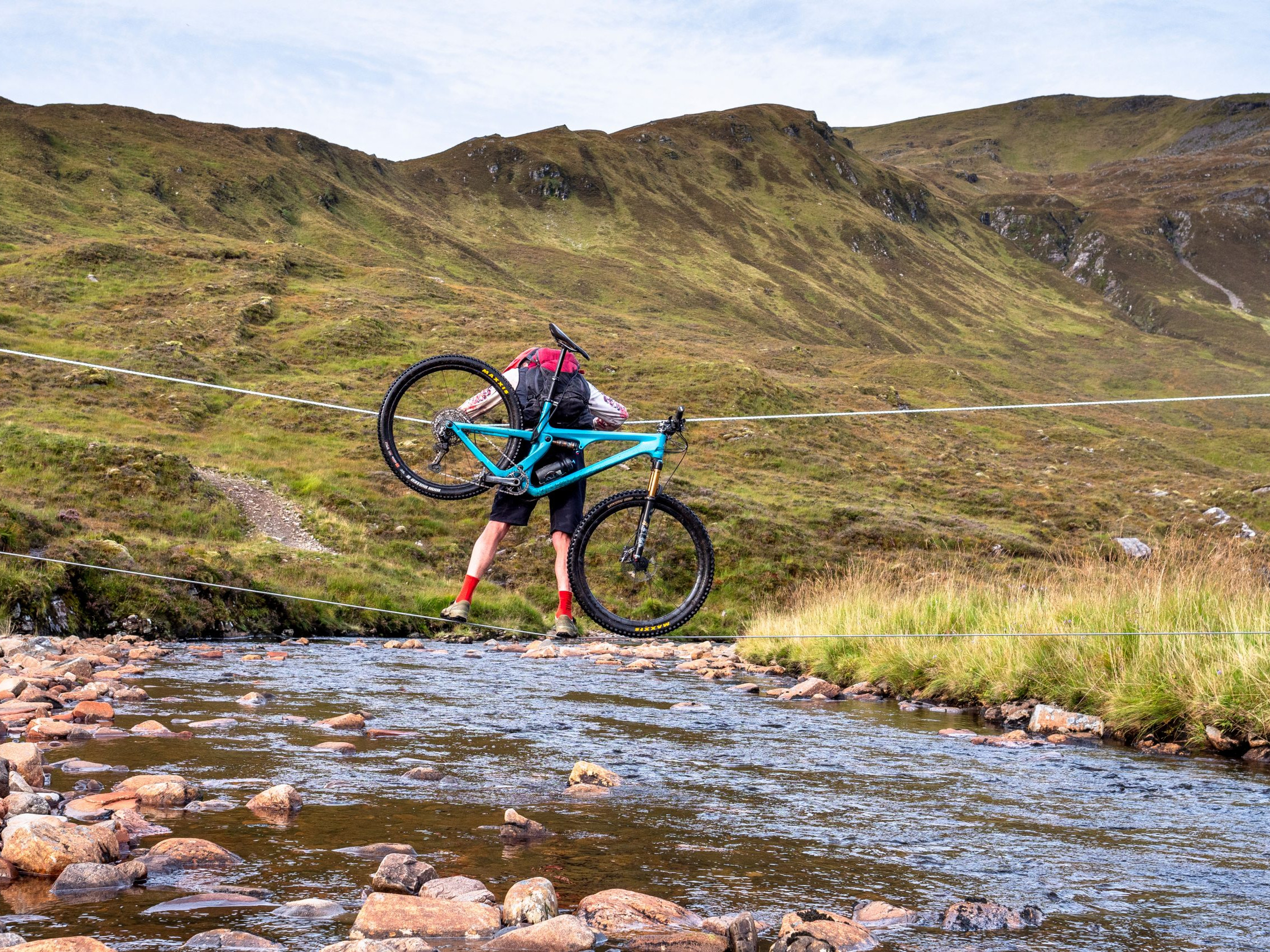 Cyclist carrying mountain bike across river on tightrope, Scottish Highlands. Adventure cycling, outdoor challenge, rural landscape. Mountain Bike United.
