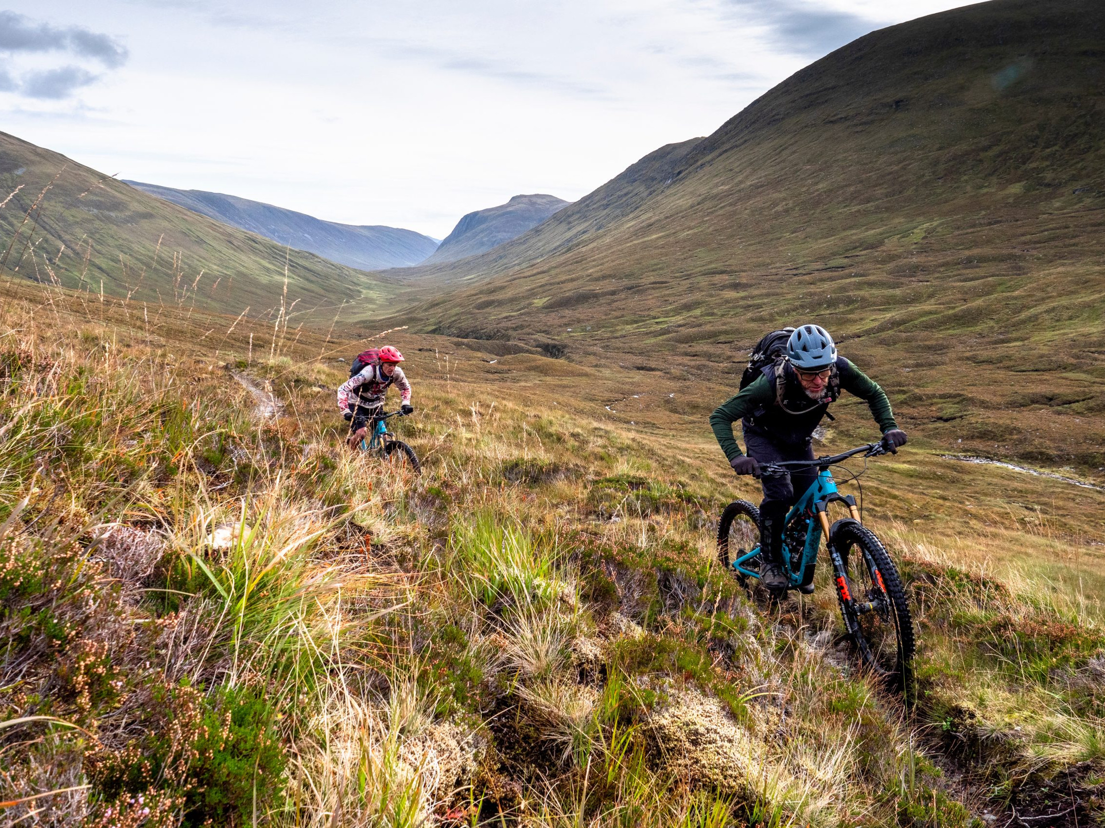 Two mountain bikers ride uphill on a grassy trail in a scenic valley, showcasing outdoor adventure and cycling terrain exploration. Mountain Bike United.