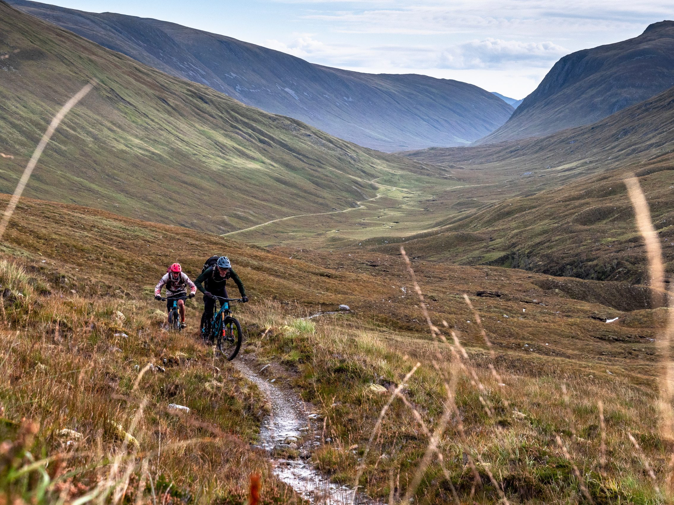 Two mountain bikers ride on a narrow trail in a vast valley landscape, ideal for outdoor biking and adventure sports enthusiasts. Mountain Bike United.