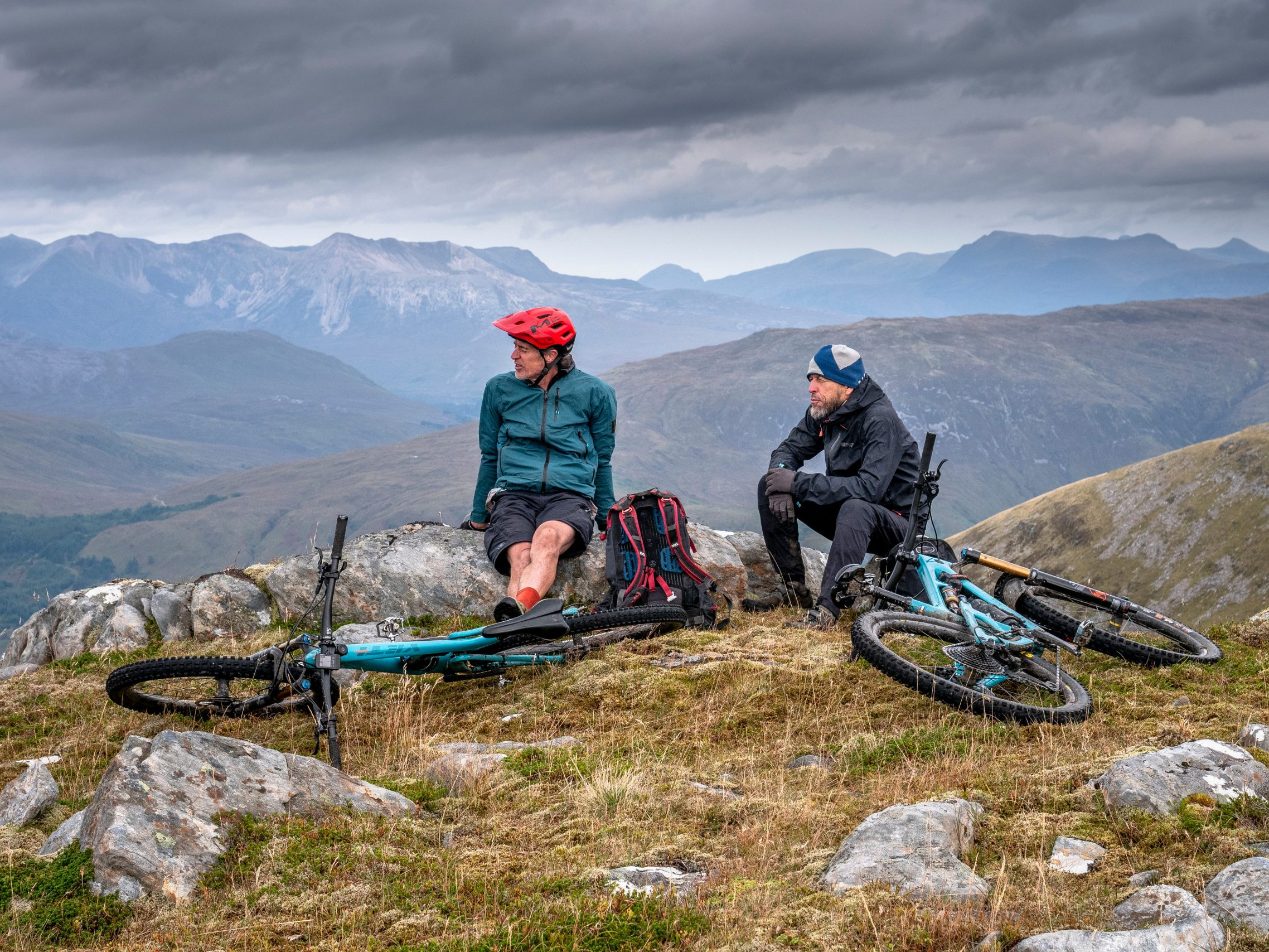 Two mountain bikers resting on a rocky hilltop with bikes, scenic mountain view, cloudy sky. Outdoor adventure, cycling, nature exploration. Mountain Bike United.