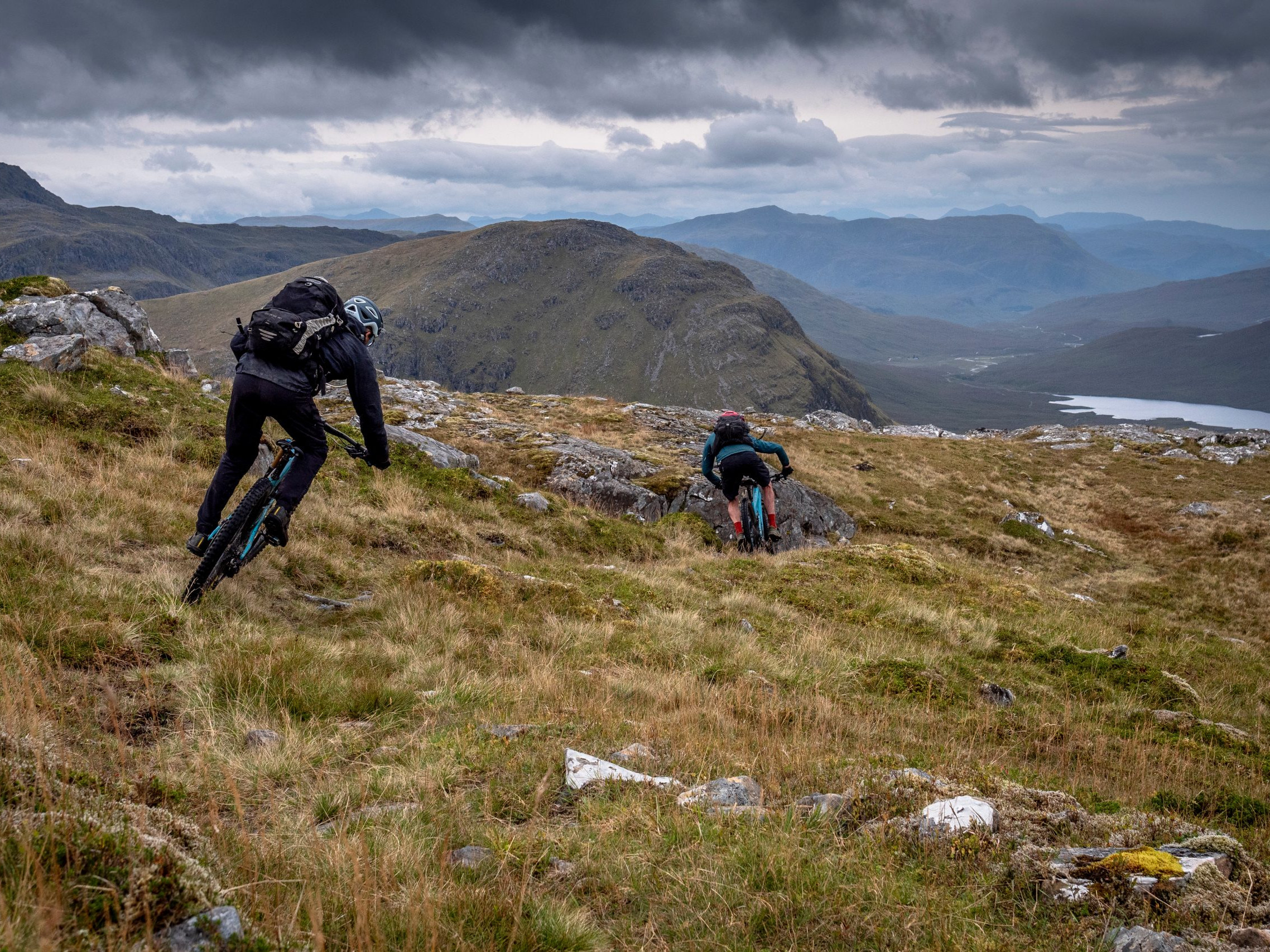 Mountain bikers riding downhill on a rugged trail in a remote, scenic mountain landscape under a cloudy sky, ideal for outdoor adventure enthusiasts. Mountain Bike United.