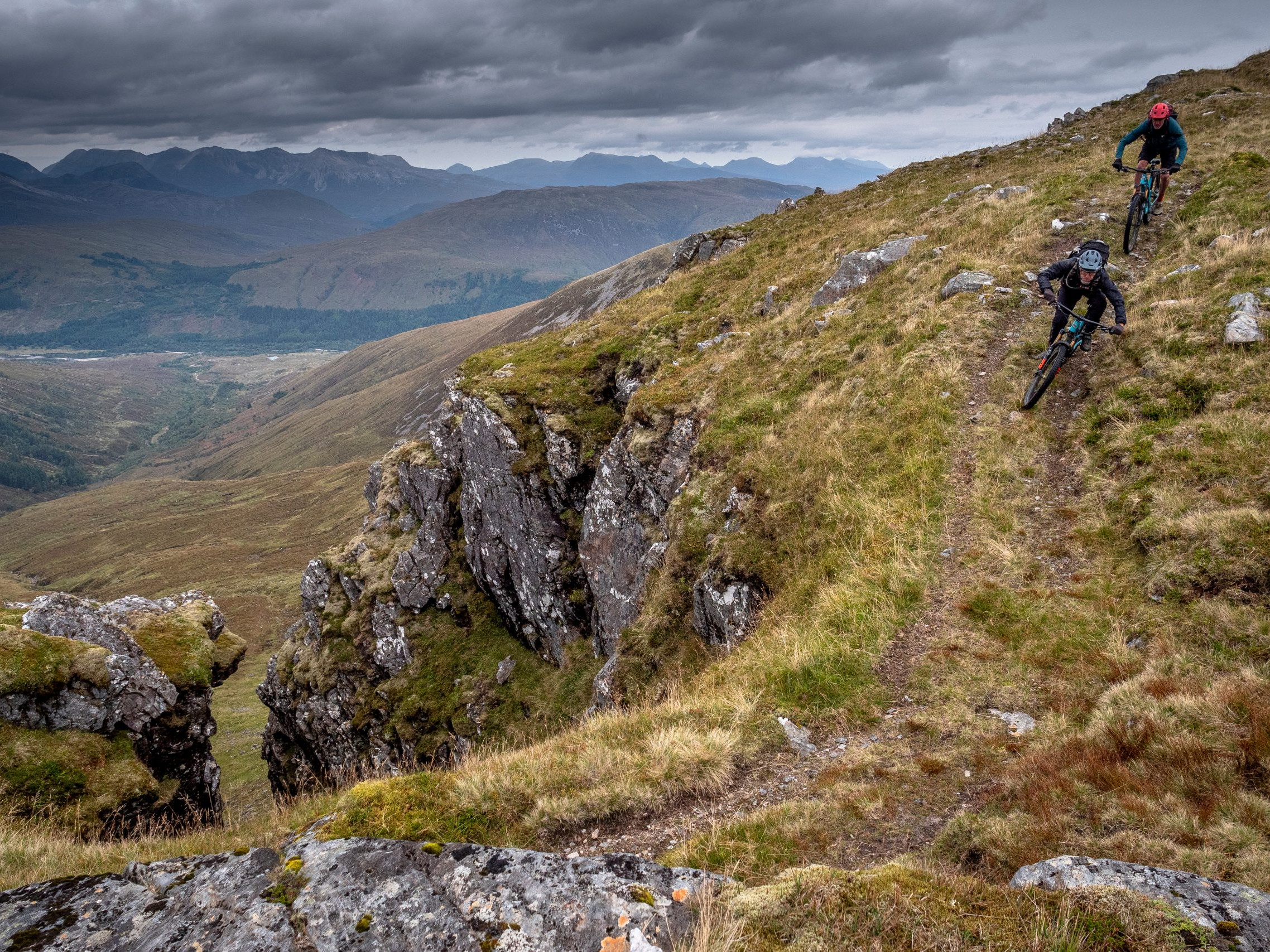 Mountain bikers descending rugged trail with scenic mountain view, overcast skies. Adventure, outdoor cycling, nature exploration in dramatic terrain. Mountain Bike United.