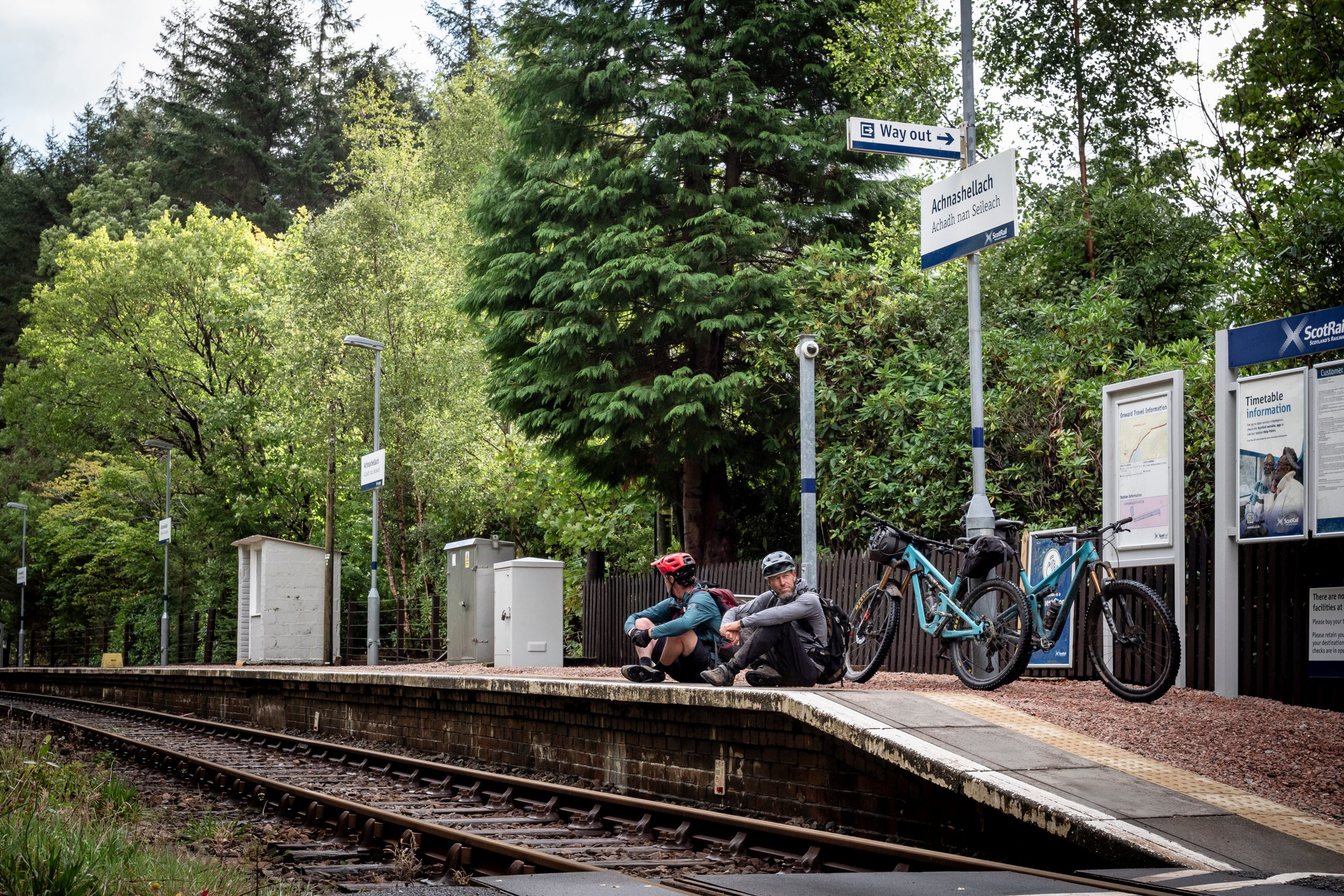 Cyclists resting at Achnashellach train station platform with bikes, surrounded by trees, Scotland. Mountain Bike United.
