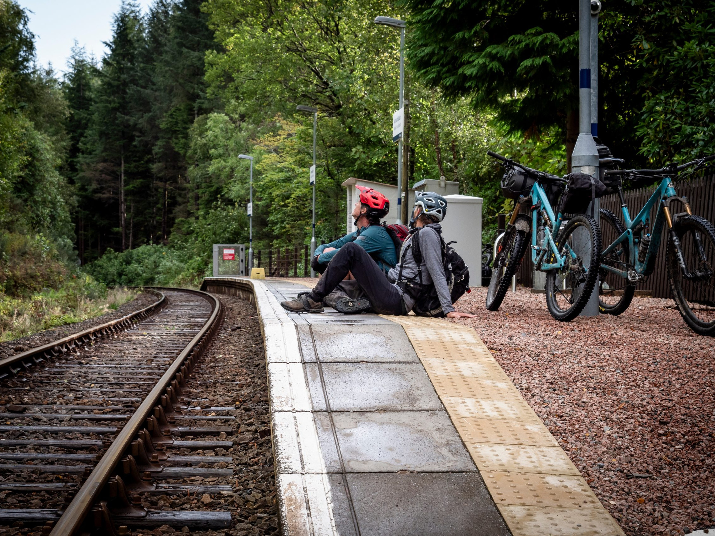 Two cyclists resting at a forest train station platform with mountain bikes, surrounded by greenery, emphasizing outdoor adventure and travel. Mountain Bike United.