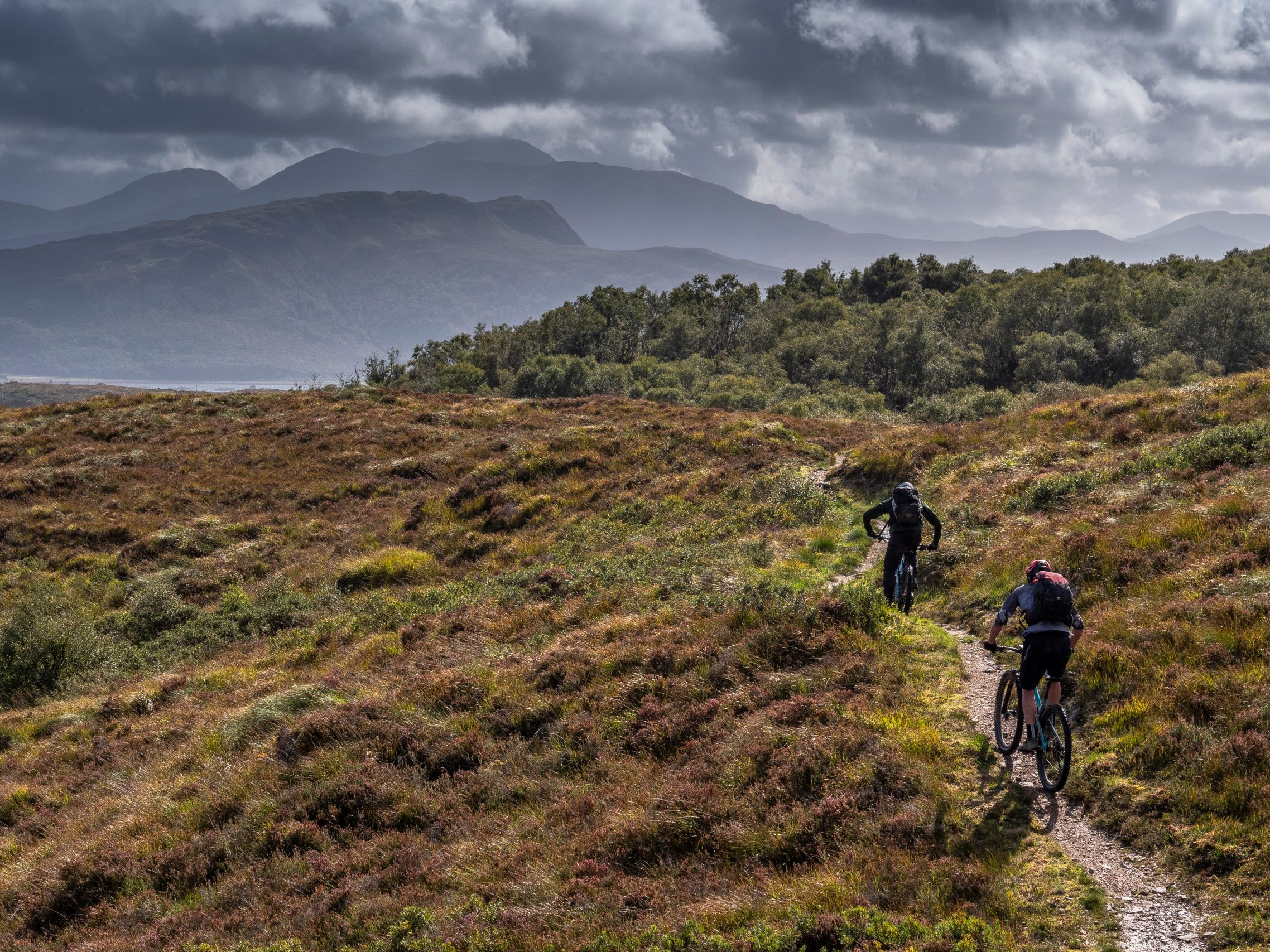 Mountain bikers on a scenic trail with dramatic skies and hills in the background, perfect for adventure and outdoor recreation enthusiasts. Mountain Bike United.