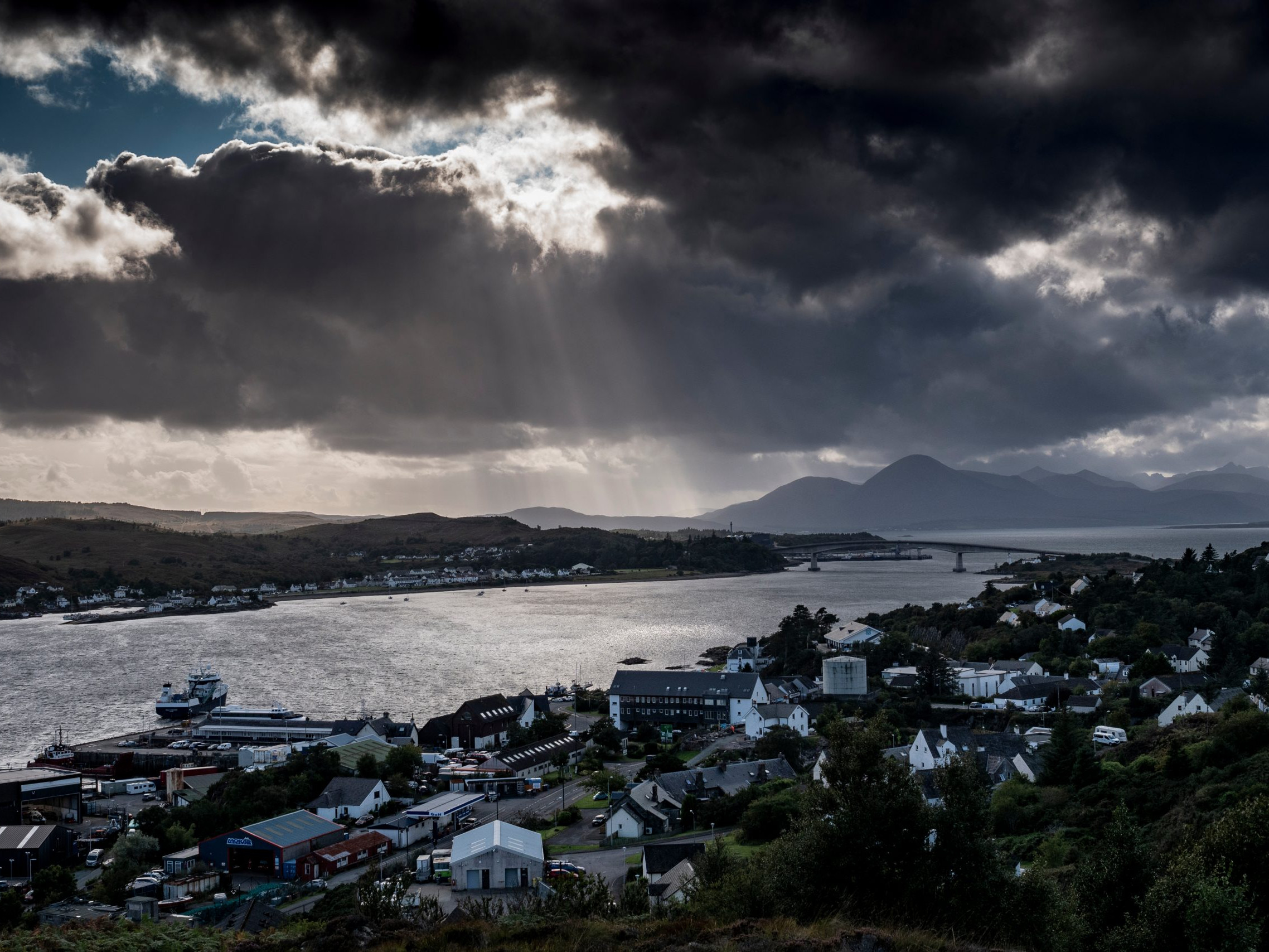 Cloudy sky over a coastal town with a bridge and mountains in the distance, highlighting dramatic weather and scenic views. Mountain Bike United.