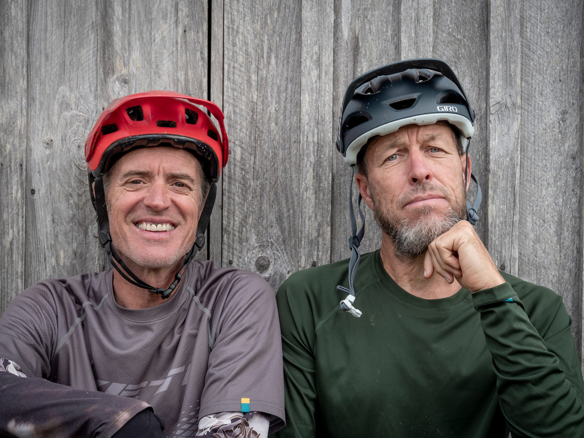 Two men wearing bike helmets stand against a wooden background. One smiles, while the other appears thoughtful. Outdoor cycling gear. Mountain Bike United.