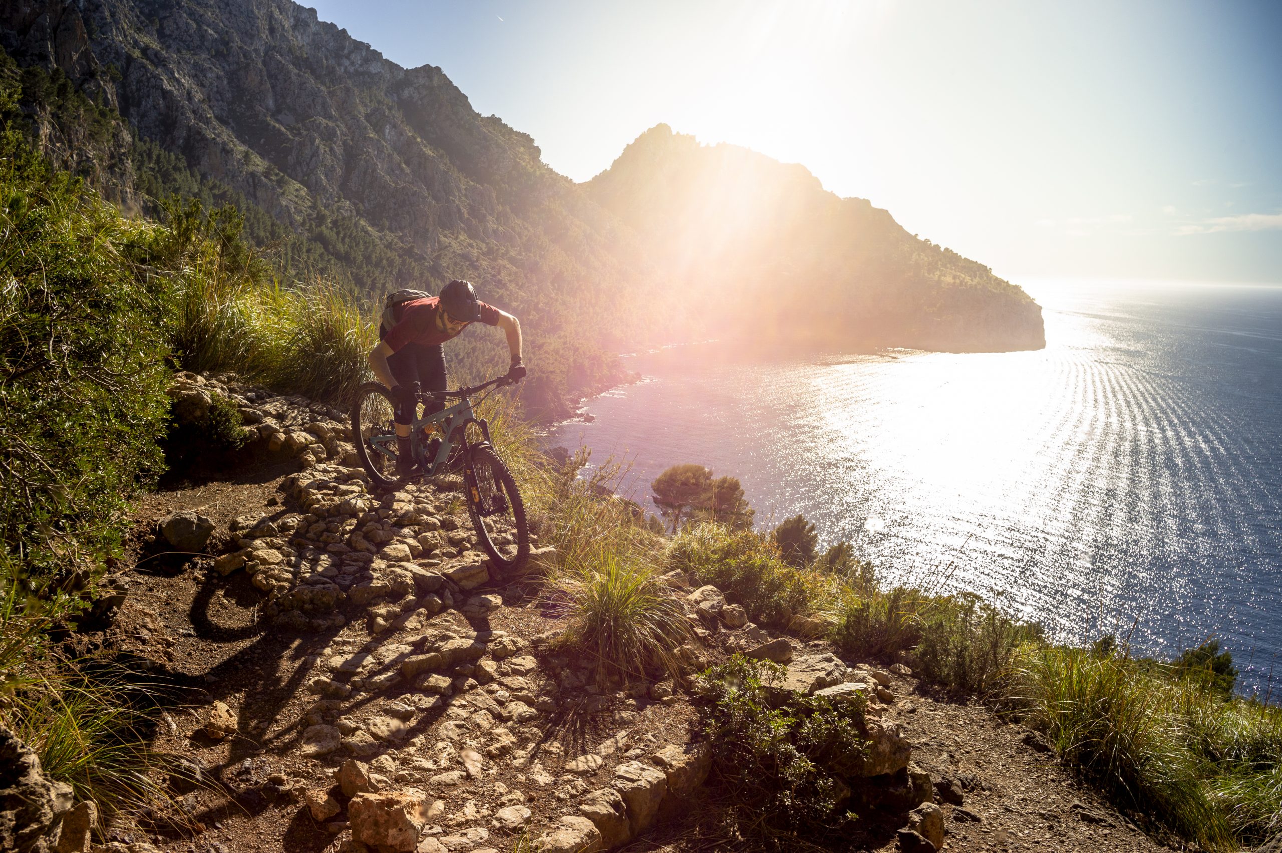 Mountain biker riding on a rocky trail by the coast, with sunlit ocean and rugged hills in background, perfect for outdoor adventure enthusiasts. Mountain Bike United.