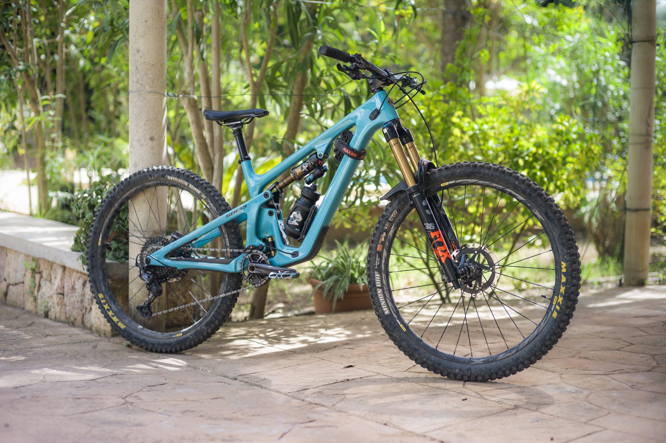 Blue mountain bike with full suspension and Maxxis tires, parked outdoors on a stone patio, surrounded by lush greenery. Mountain Bike United.