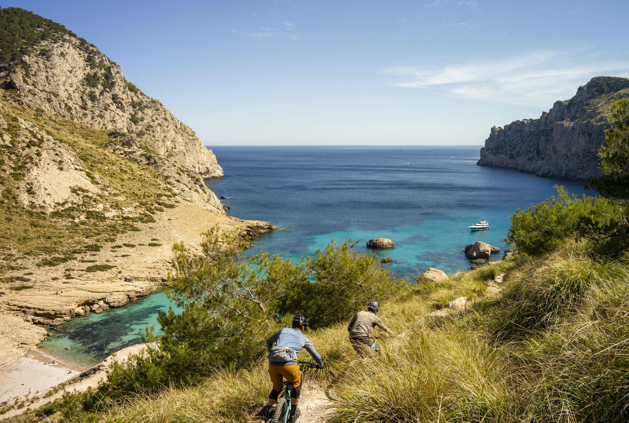 Mountain bikers ride on a coastal trail overlooking a stunning blue sea with cliffs and a boat, ideal for outdoor adventure and scenic views. Mountain Bike United.