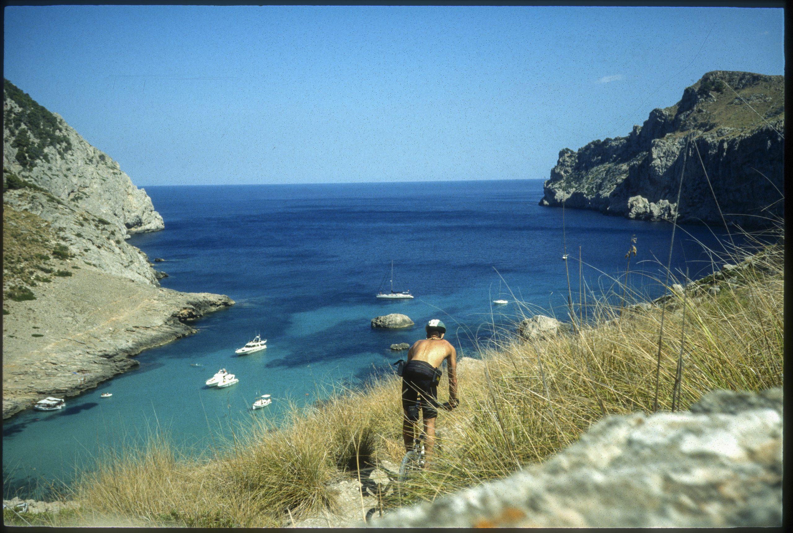 Cyclist overlooking Mediterranean bay with rocky cliffs and yachts, Majorca coast, clear blue water, adventure cycling, scenic sea view, outdoor exploration. Mountain Bike United.