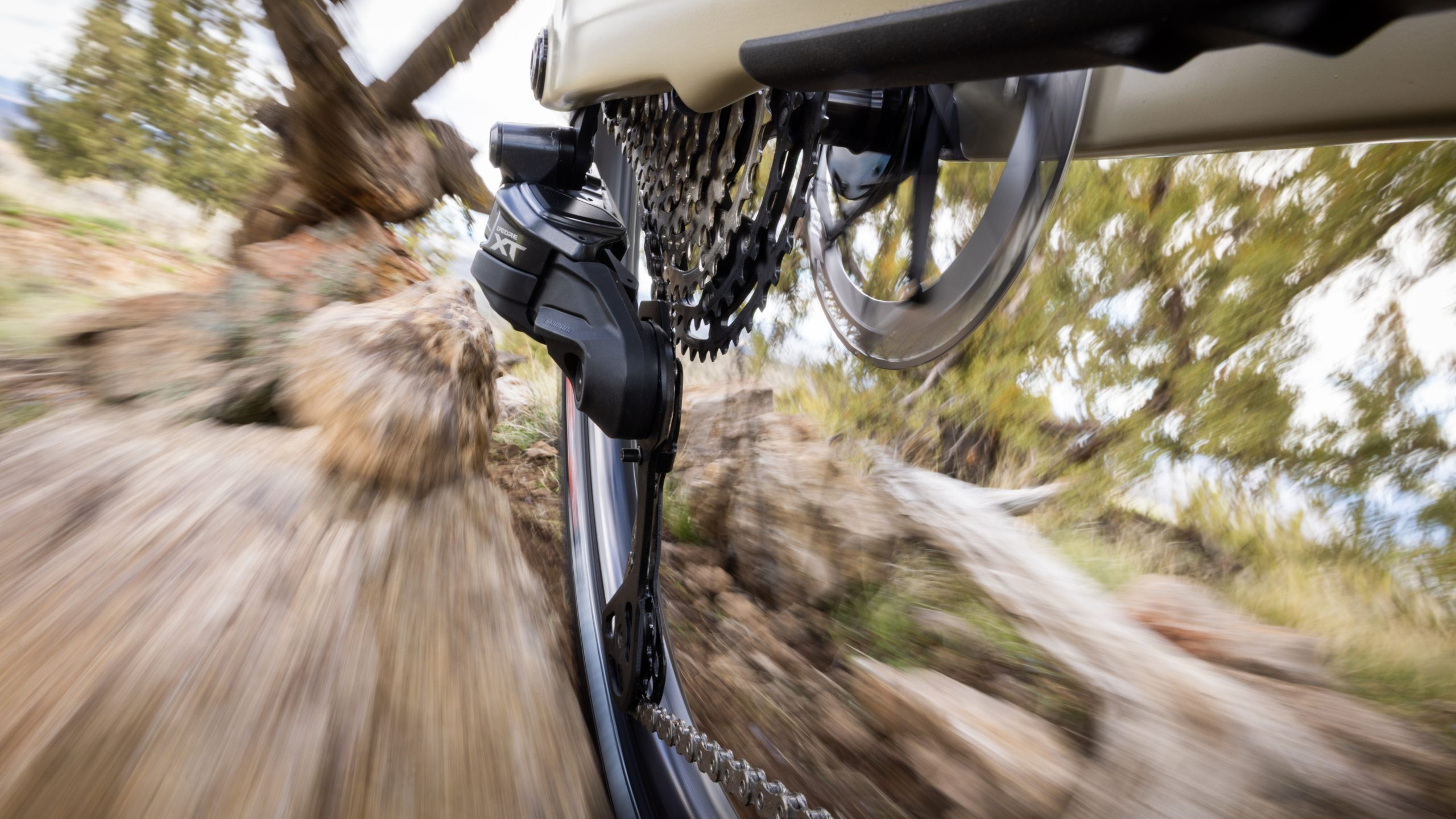 Mountain bike gear system in motion on a rocky trail, featuring Shimano XT derailleur and spinning wheel, showcasing off-road cycling dynamics. Mountain Bike United.