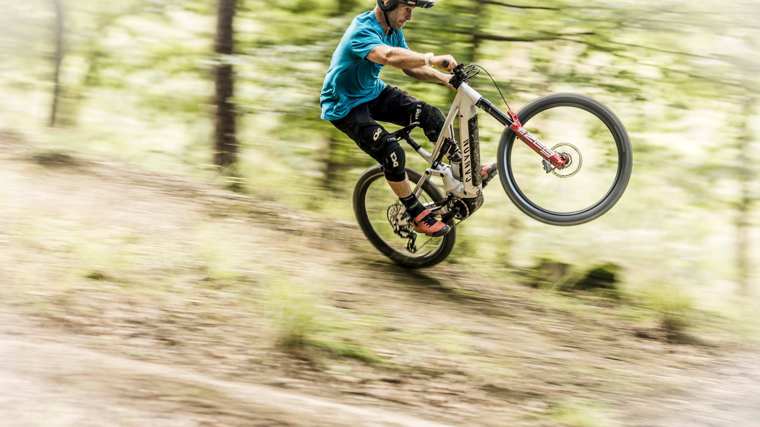 Mountain biker performing wheelie in forest, wearing safety gear, showcasing bike control and agility on woodland trail. Mountain Bike United.