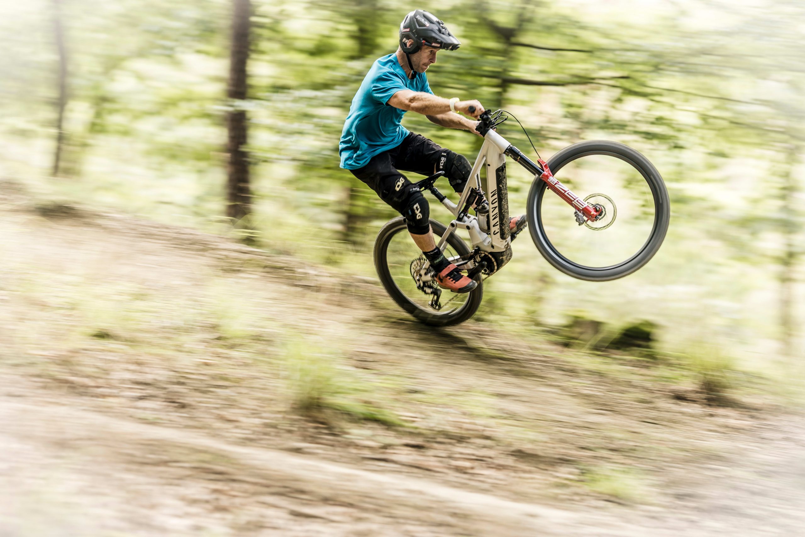 Mountain biker performing wheelie in forest, wearing safety gear, showcasing bike control and agility on woodland trail. Mountain Bike United.