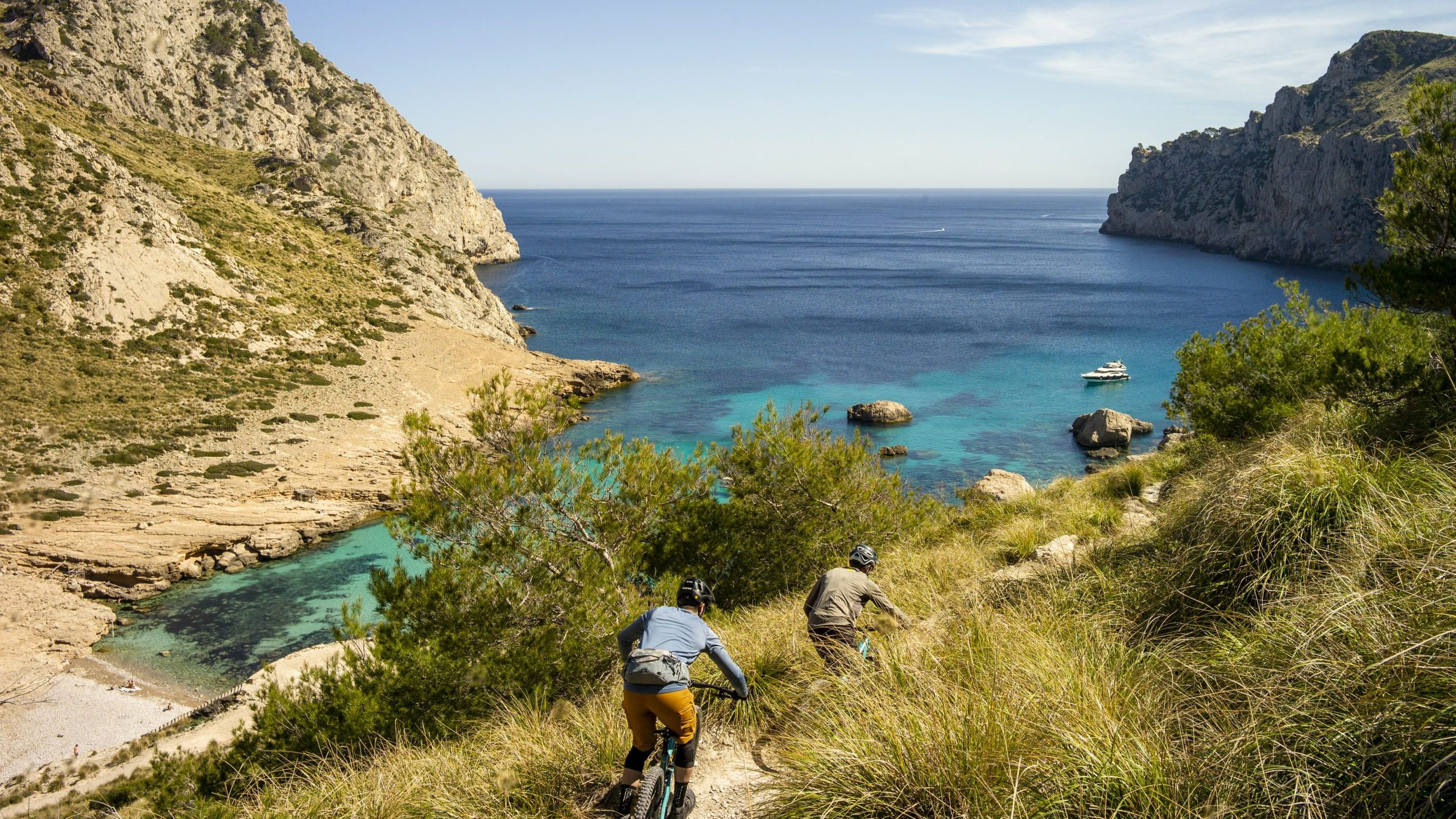 Cyclists biking down a coastal trail with lush cliffs and turquoise waters, and a yacht floating in the Mediterranean Sea in the background. Mountain Bike United.