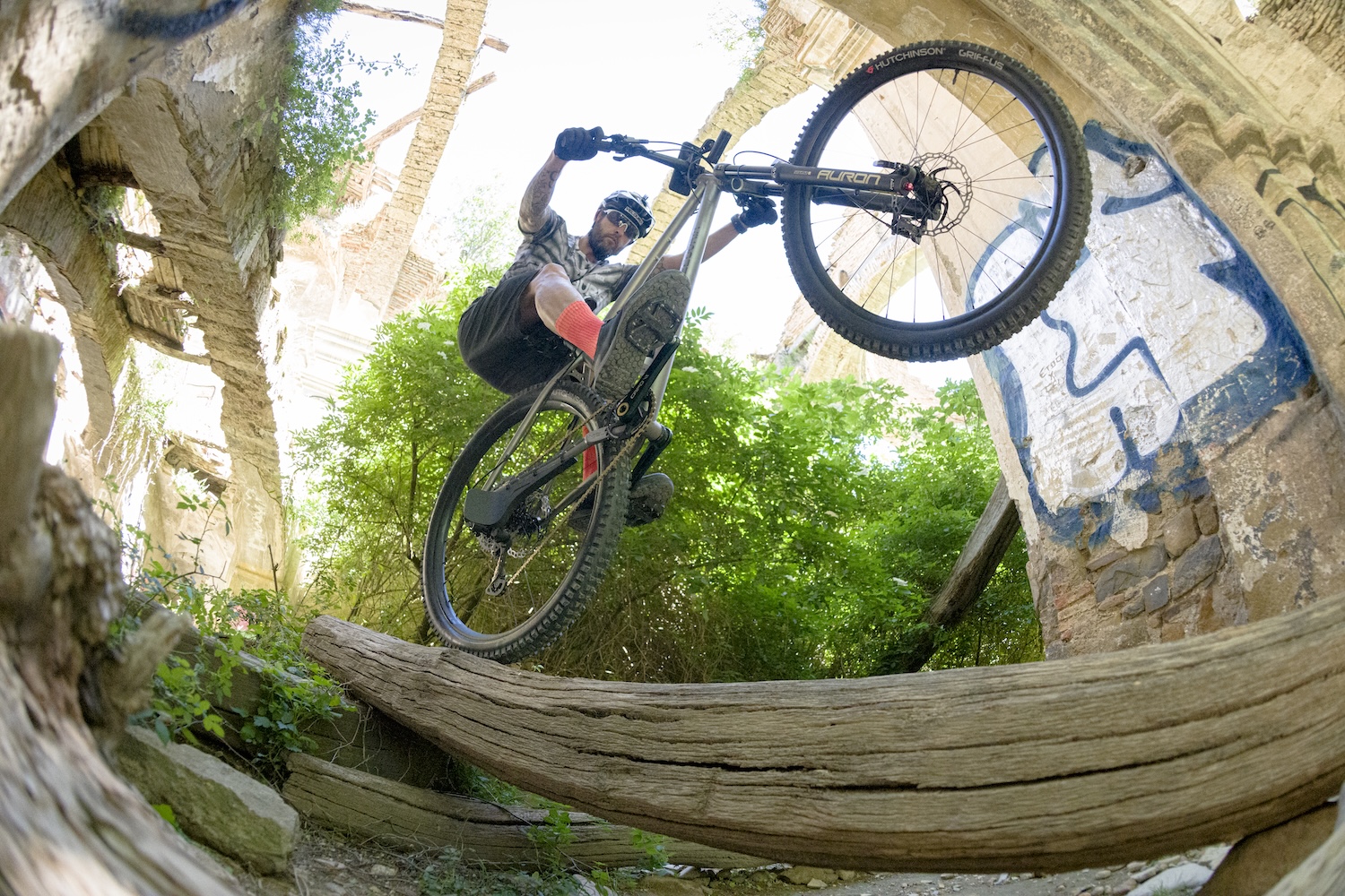 Mountain biker performing a jump over a log in an abandoned building, surrounded by greenery and graffiti, showcasing extreme biking skills. Mountain Bike United.