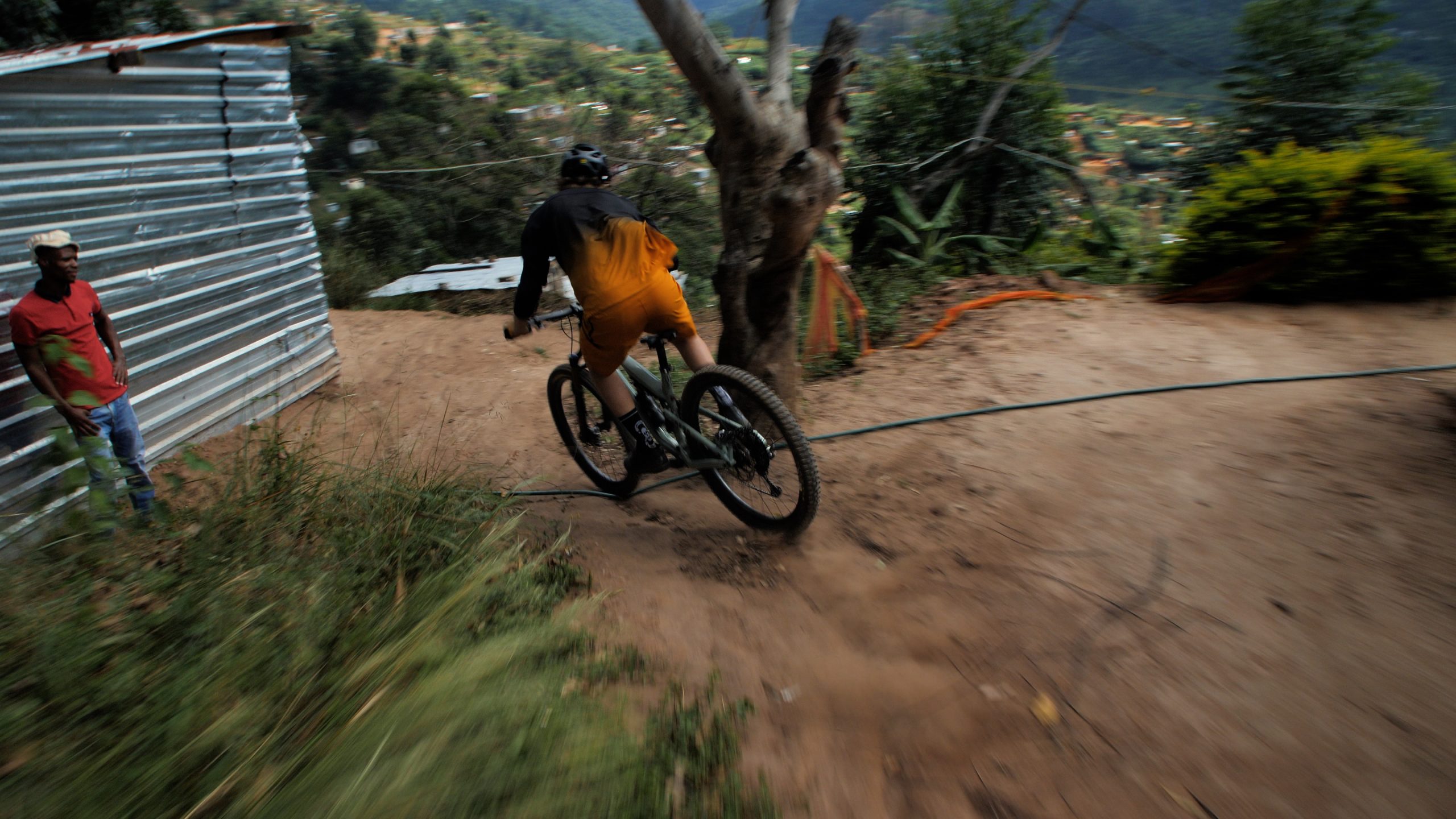 Mountain biker turning on dirt path, wearing helmet and sports gear. Another person stands nearby; rural setting with trees and corrugated metal structure. Mountain Bike United.