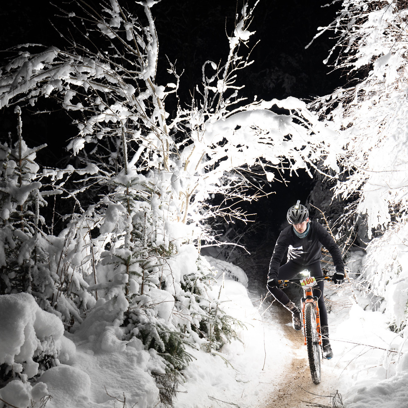 Cyclist riding mountain bike through snowy forest trail at night, wearing helmet and winter gear, highlighting winter biking adventure. Mountain Bike United.