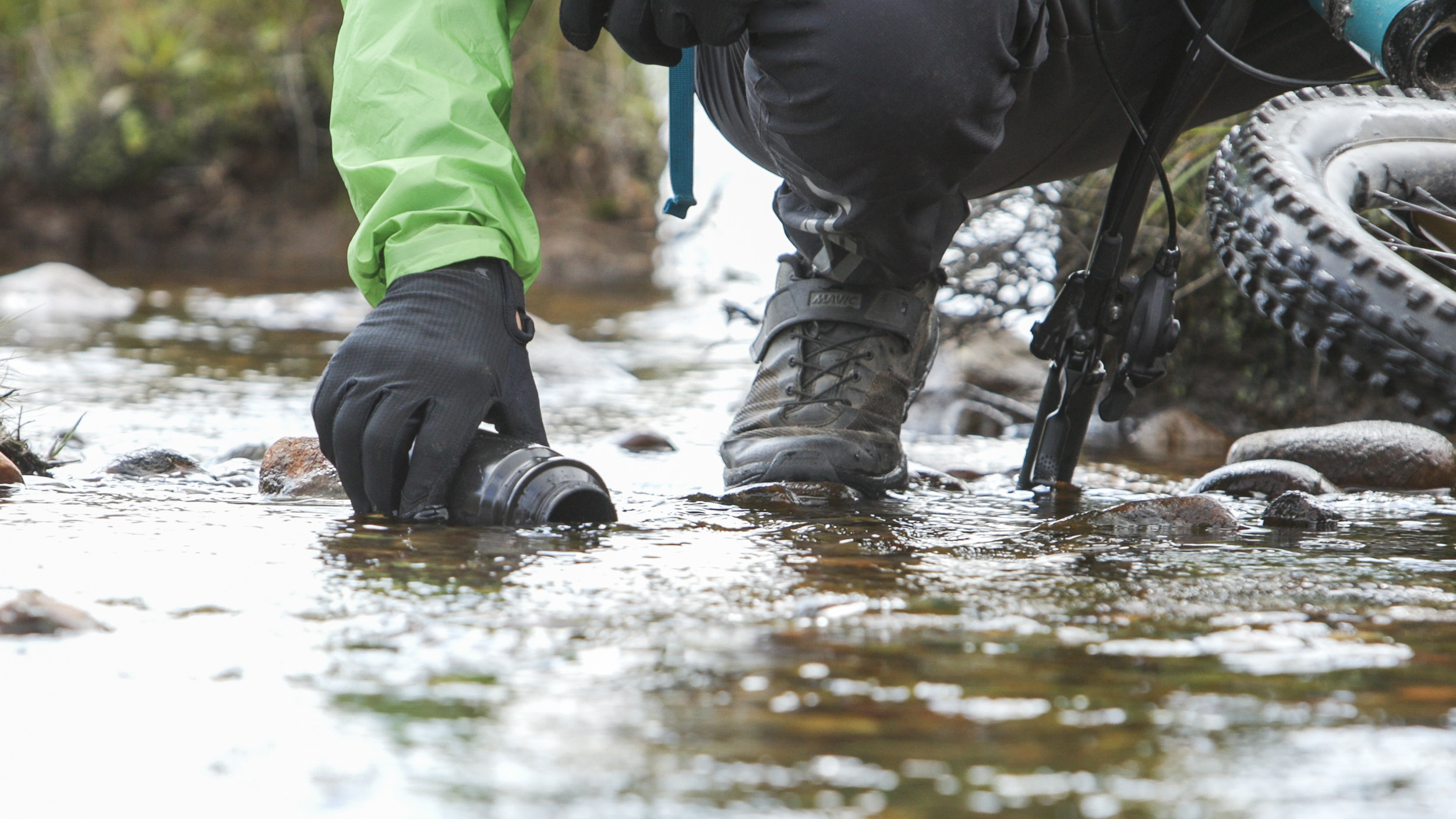 Cyclist in green jacket kneeling by stream, filling cup with water. Dan Milner, A Single Drop concept in outdoor adventure. Mountain Bike United.