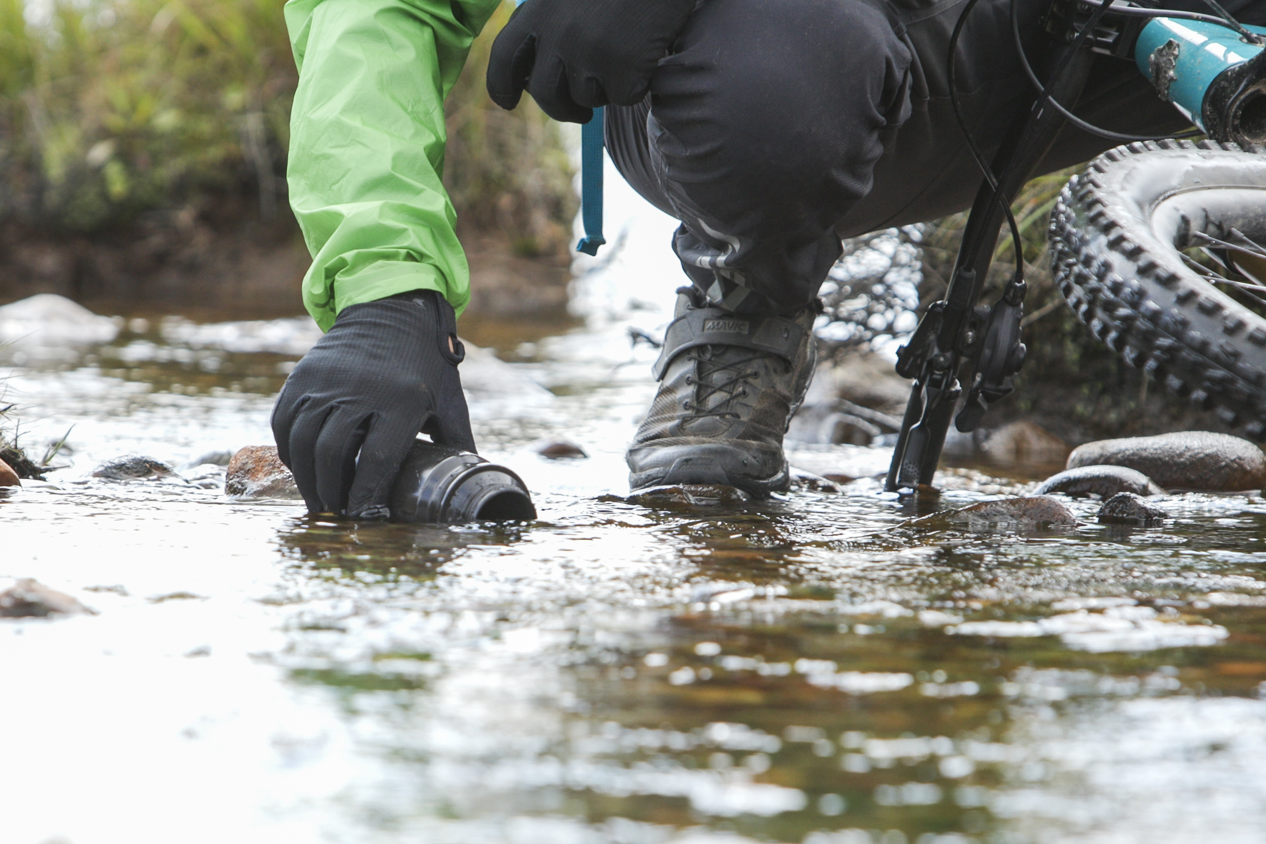 Cyclist in green jacket kneeling by stream, filling cup with water. Dan Milner, A Single Drop concept in outdoor adventure. Mountain Bike United.
