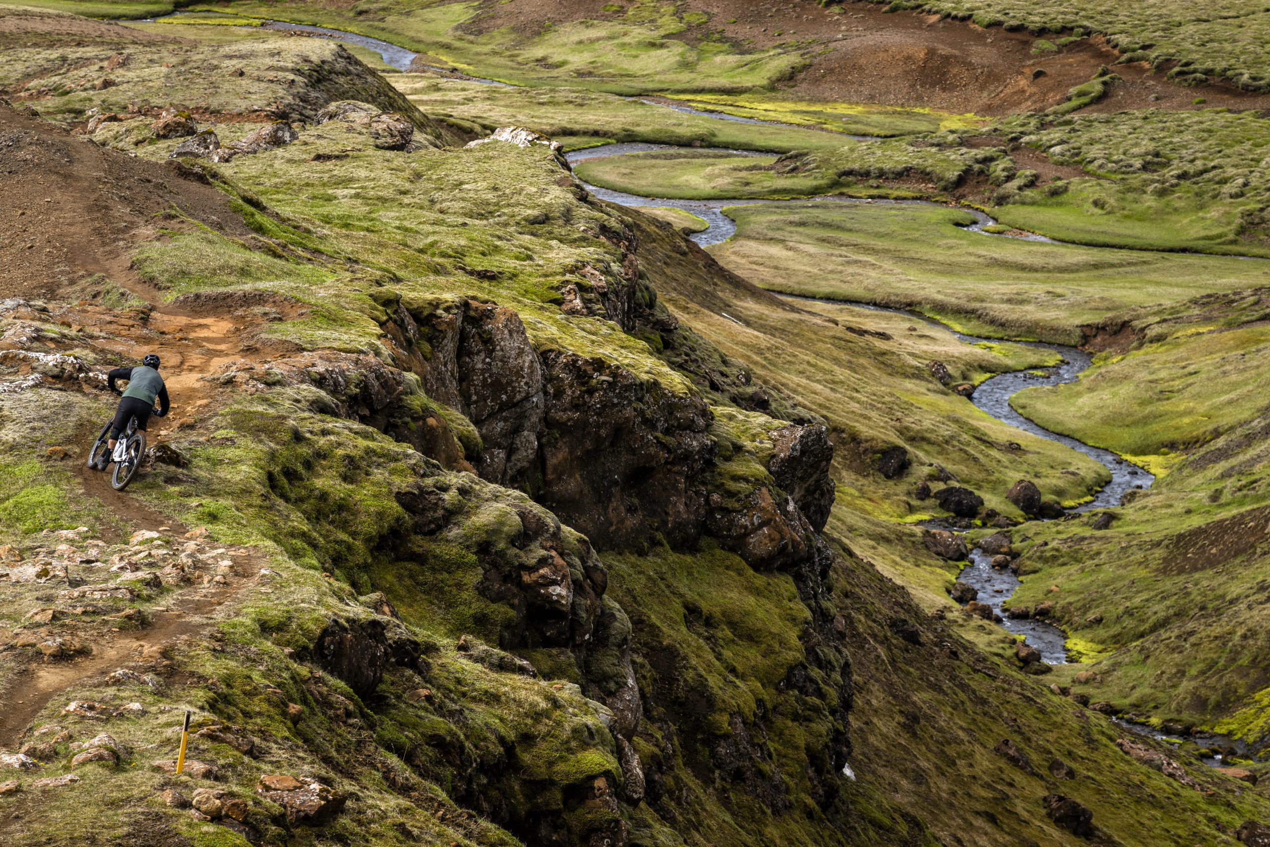 Mountain biker on rugged trail, green landscape, resembling Andrew Neethling's Journey to The Centre of The Earth adventure scene. Mountain Bike United.