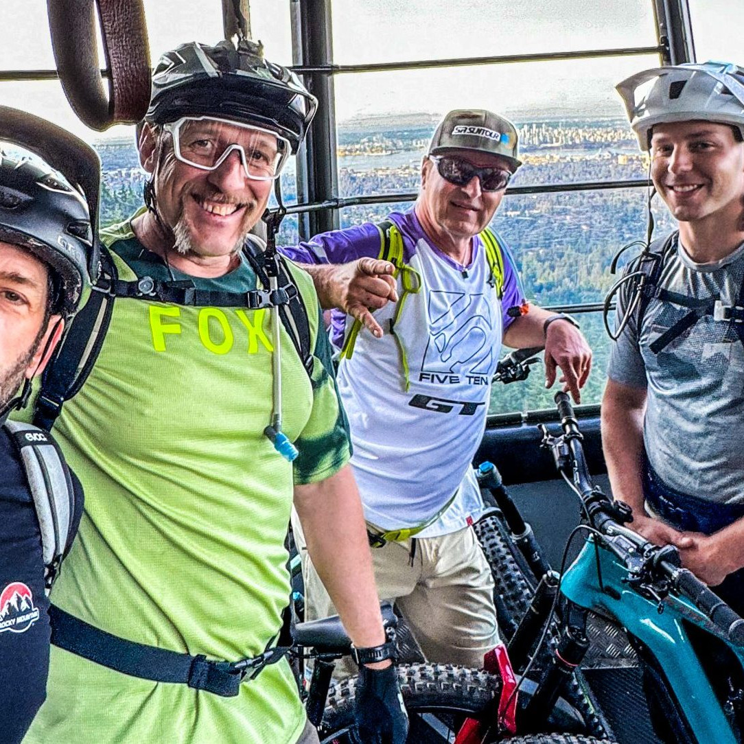 Four mountain bikers with gear inside a gondola, enjoying a scenic view before a downhill ride. Relevant to Hans Rey's biking adventures. Mountain Bike United.