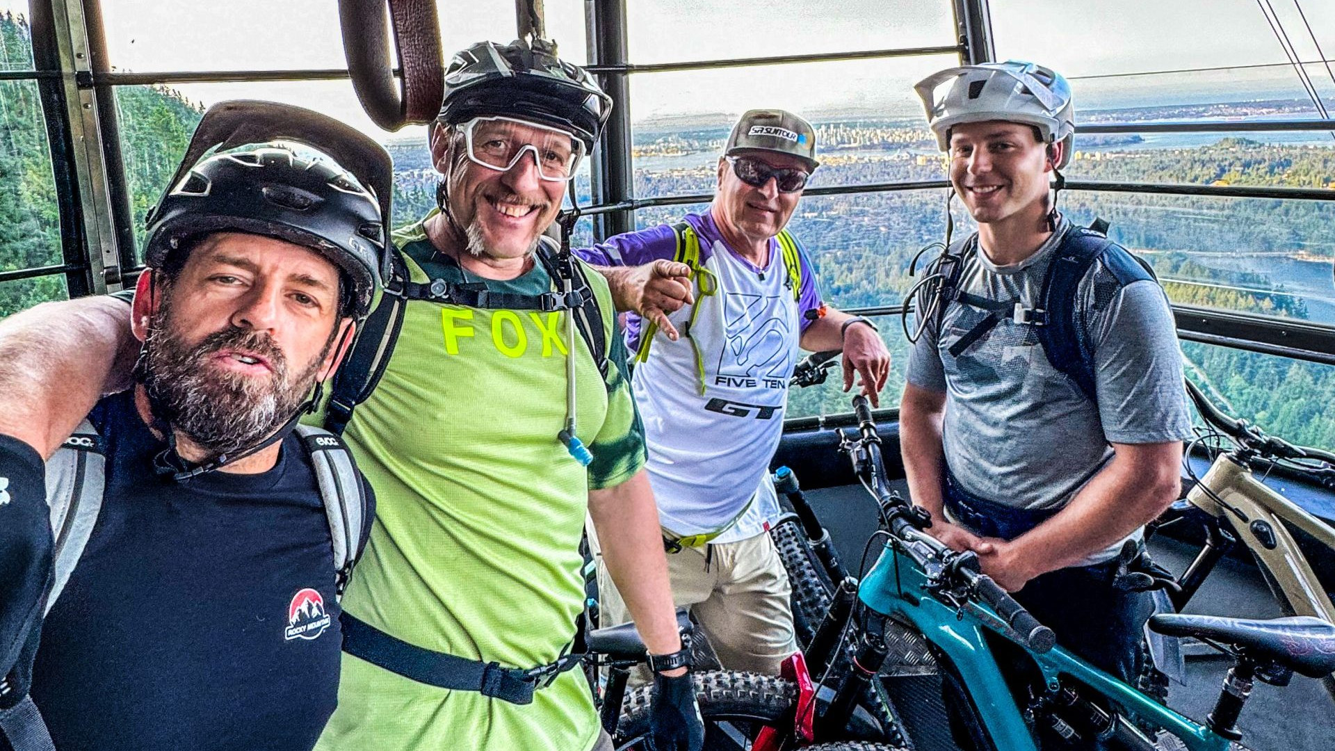 Four mountain bikers with gear inside a gondola, enjoying a scenic view before a downhill ride. Relevant to Hans Rey's biking adventures. Mountain Bike United.