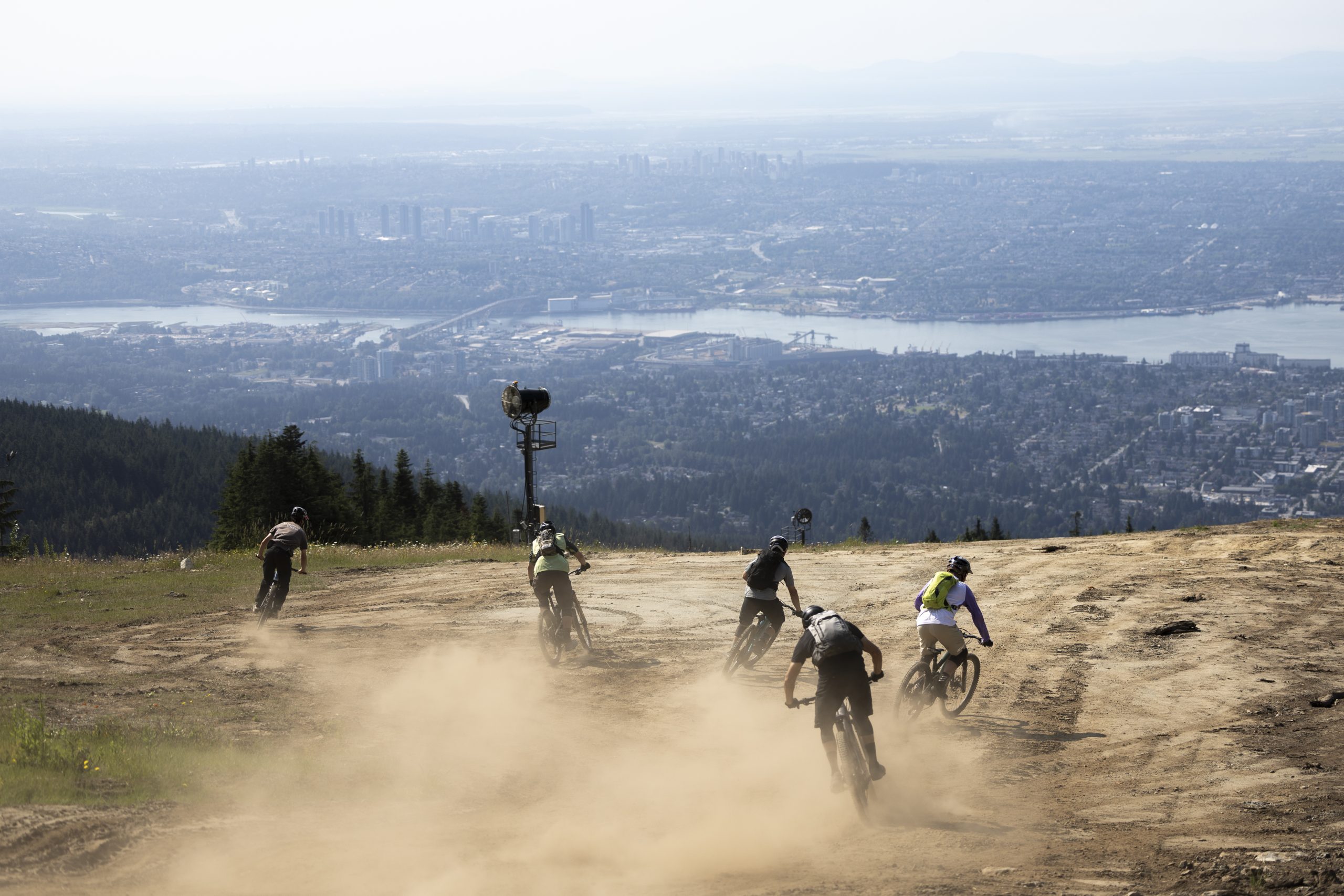 Mountain bikers ride downhill on a dusty trail with cityscape views, related to Hans Rey adventure cycling and scenic biking routes. Mountain Bike United.