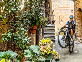 Cyclist performs a wheelie in an Umbrian alley, showcasing the adventure theme of Once Upon A Ride In Umbria with Hans Rey. Mountain Bike United.