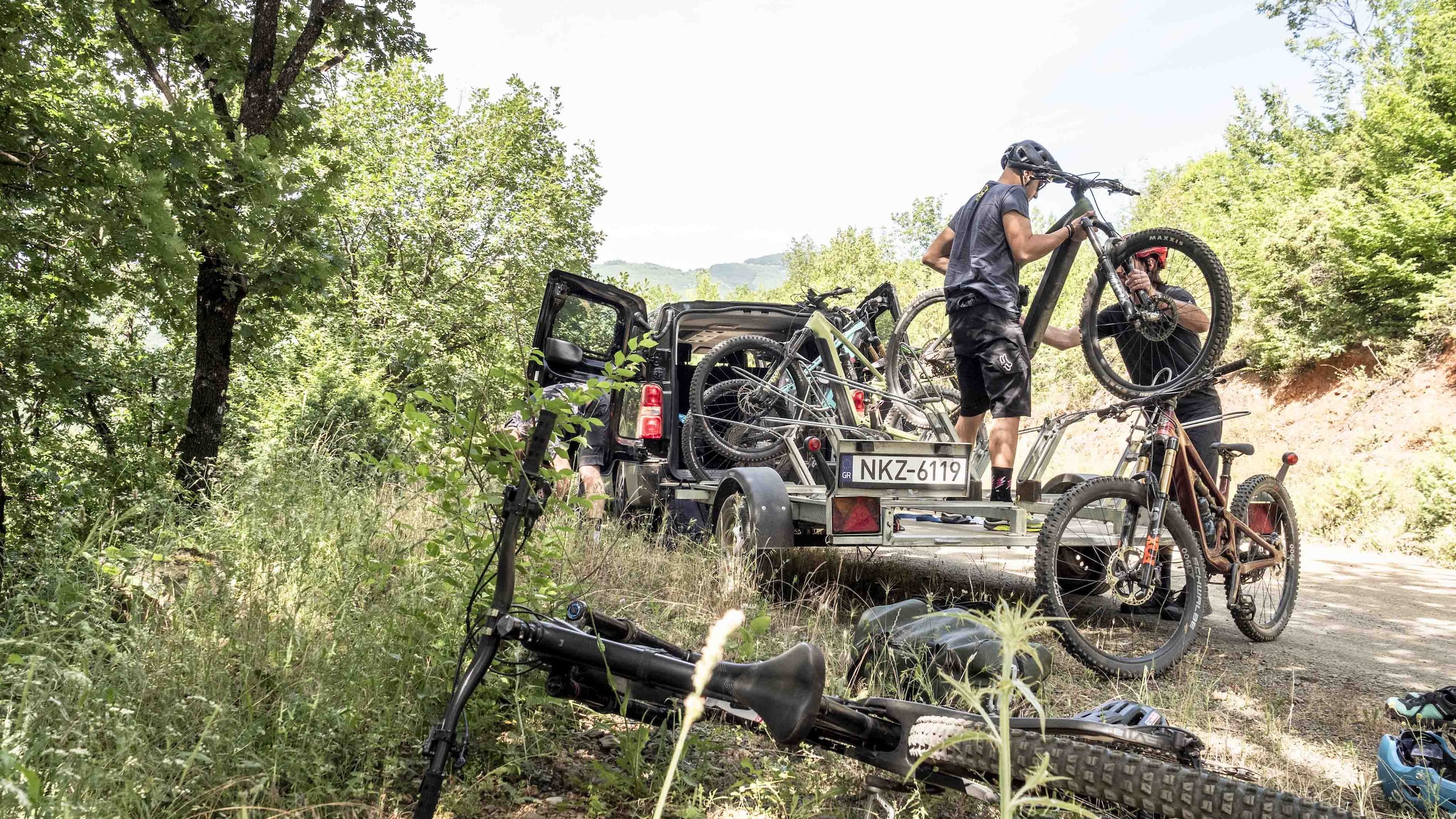 Loading mountain bikes onto a trailer in a forest, with people wearing helmets. Outdoor biking adventure, SUV, nature, and cycling equipment visible. Mountain Bike United. Digital solutions for trail organisations.