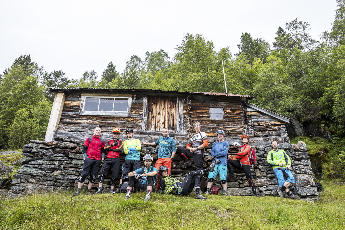 Mountain bike community app group of bikers in gear posing in front of a rustic cabin, showcasing outdoor adventure and biking camaraderie. Mountain Bike United.