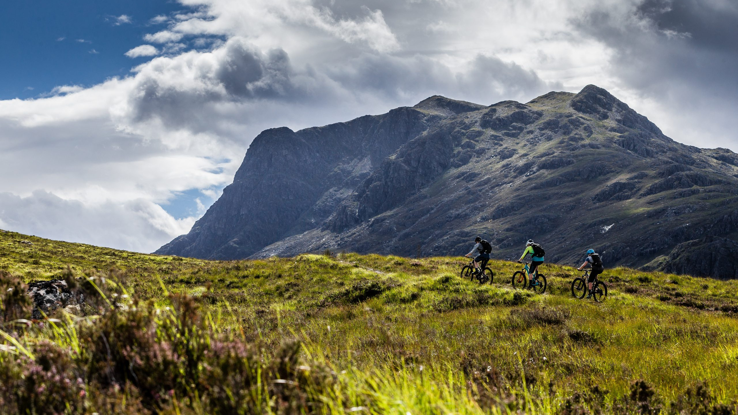 Mountain biking scene with three cyclists riding through lush green hills against dramatic mountain and cloudy sky background, outdoor adventure exploration. Mountain Bike United.