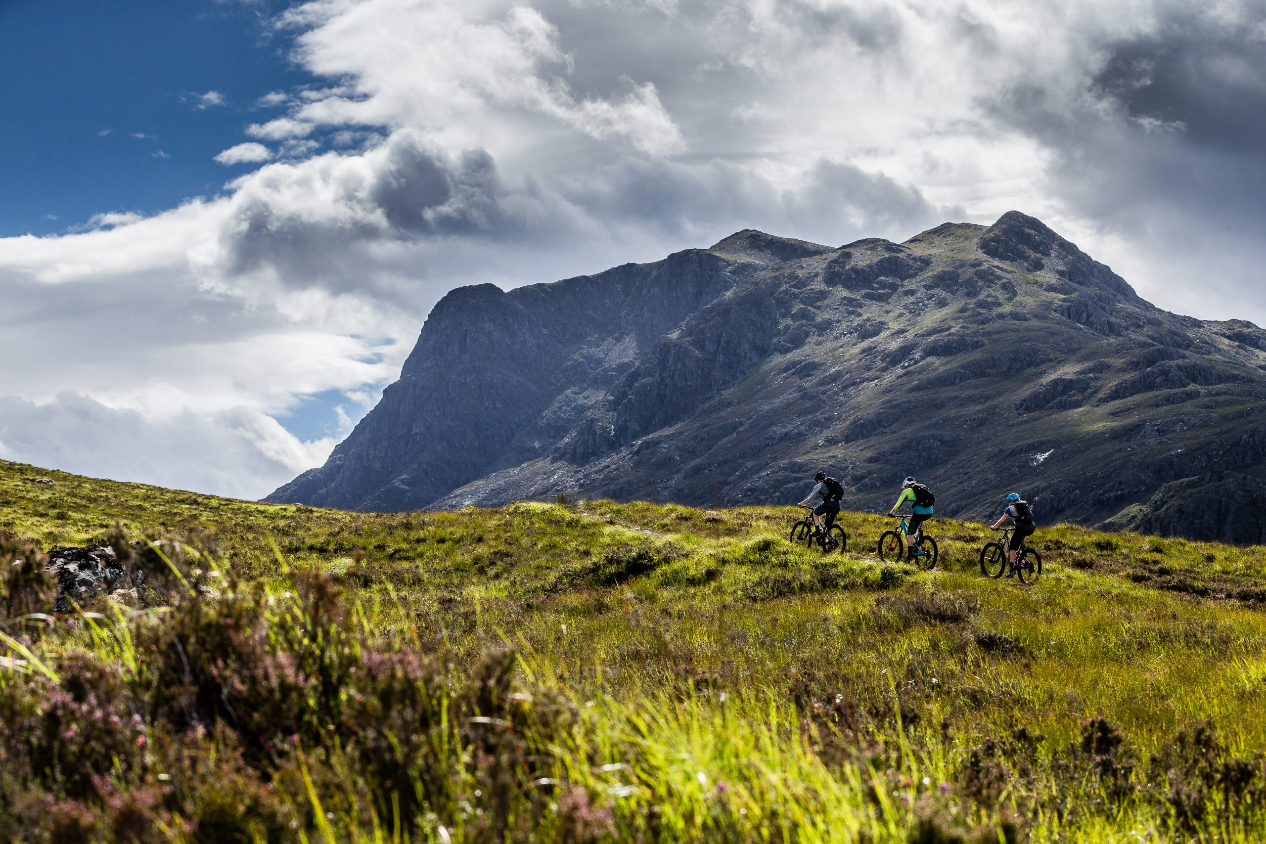 Mountain biking scene with three cyclists riding through lush green hills against dramatic mountain and cloudy sky background, outdoor adventure exploration. Mountain Bike United.