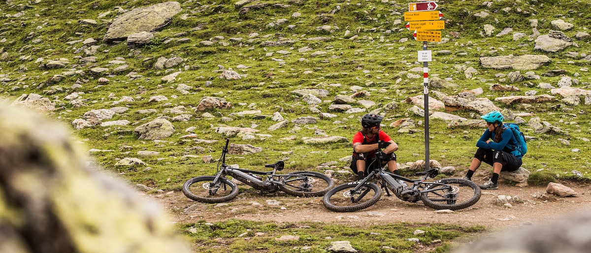 E-mountain bikers resting with bikes, using digital tools for navigation at a signpost in a rocky landscape. Mountain Bike United.