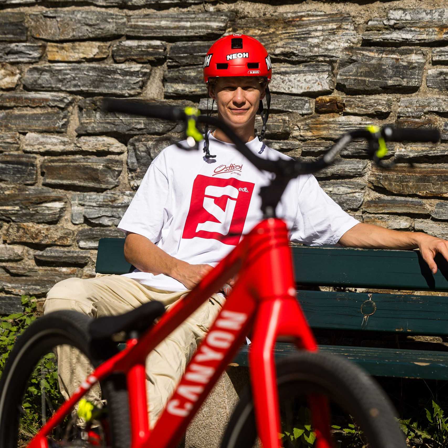 Gabriel Wibmer sitting on a bench with a red Canyon bike in the foreground, wearing a helmet and white shirt, against a stone wall. Mountain Bike United.