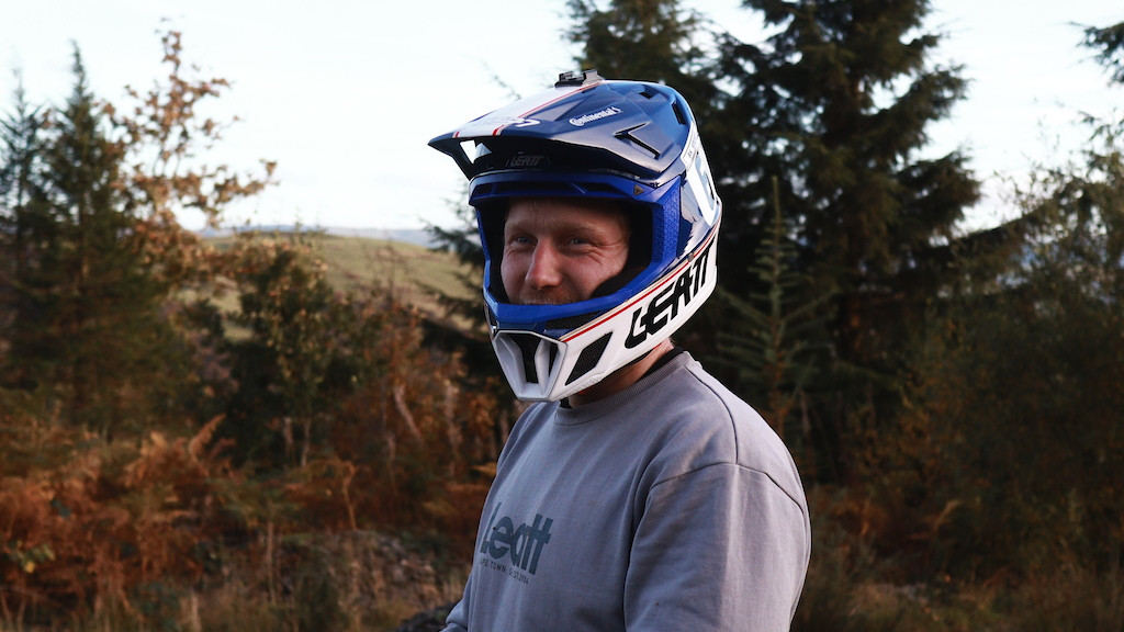 Mountain biker in a blue helmet and gray shirt holding bike handlebars, standing in a forested trail setting, ready for off-road cycling. Mountain Bike United.