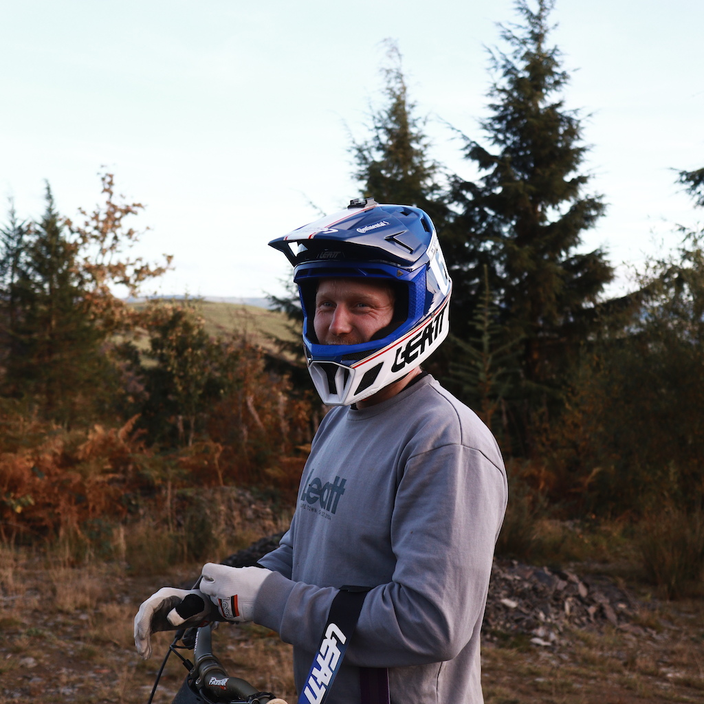 Mountain biker in a blue helmet and gray shirt holding bike handlebars, standing in a forested trail setting, ready for off-road cycling. Mountain Bike United.