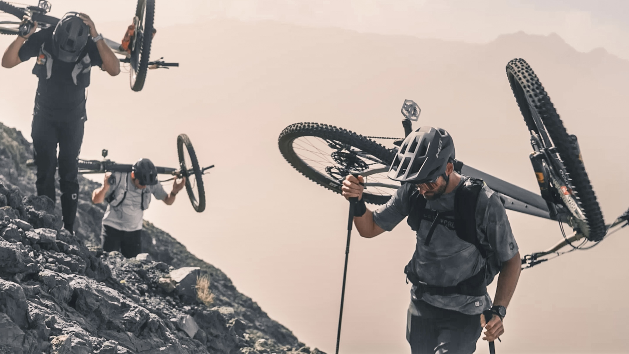 Mountain bikers carrying bikes uphill on rocky terrain, wearing helmets and gear, showcasing extreme cycling adventure and endurance in rugged landscape. Mountain Bike United.