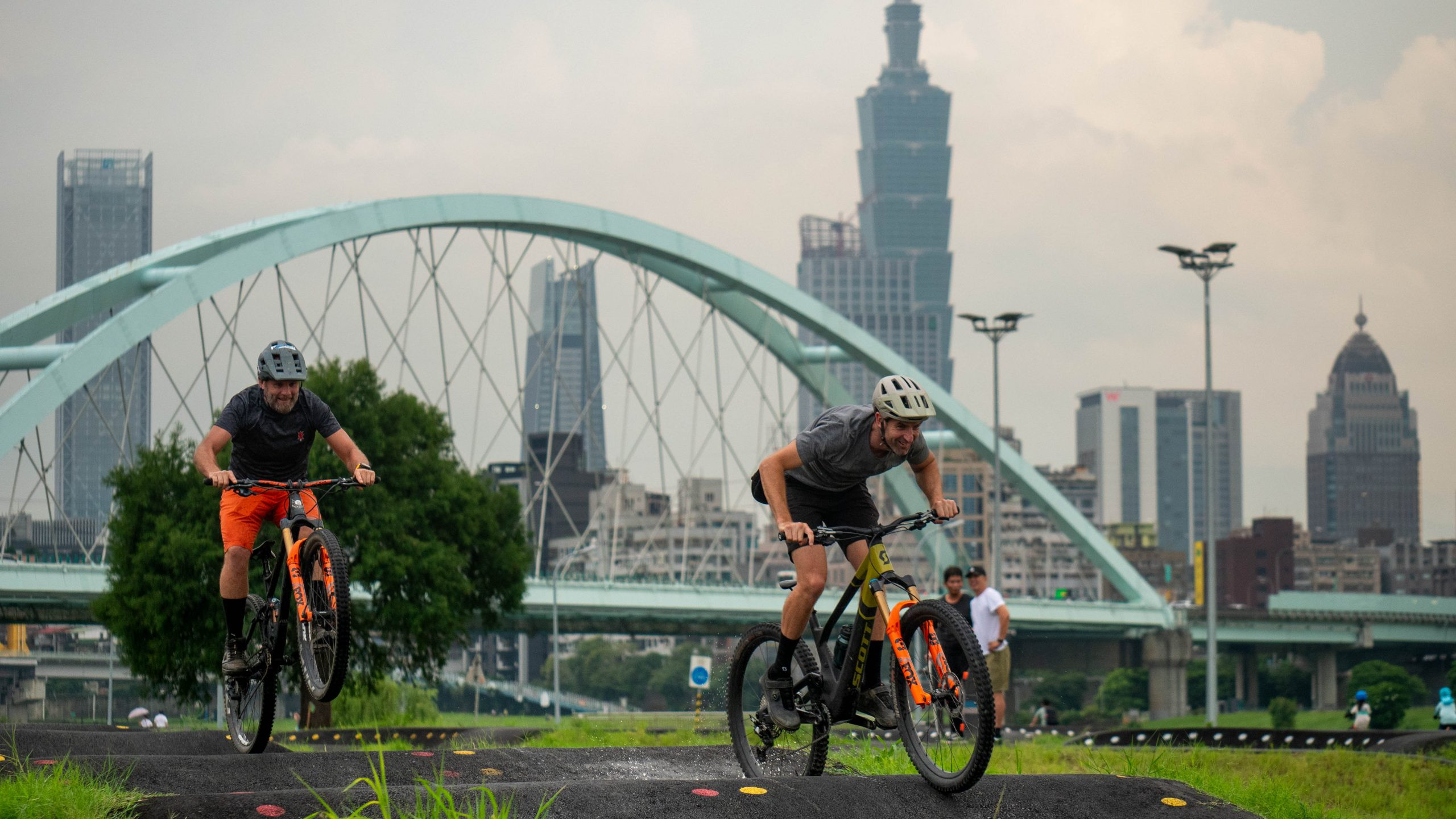 Cyclists on mountain bikes riding a pump track with cityscape backdrop, featuring a prominent arch bridge. Keywords: Andrew Neethling Lifting The Lid. Mountain Bike United.