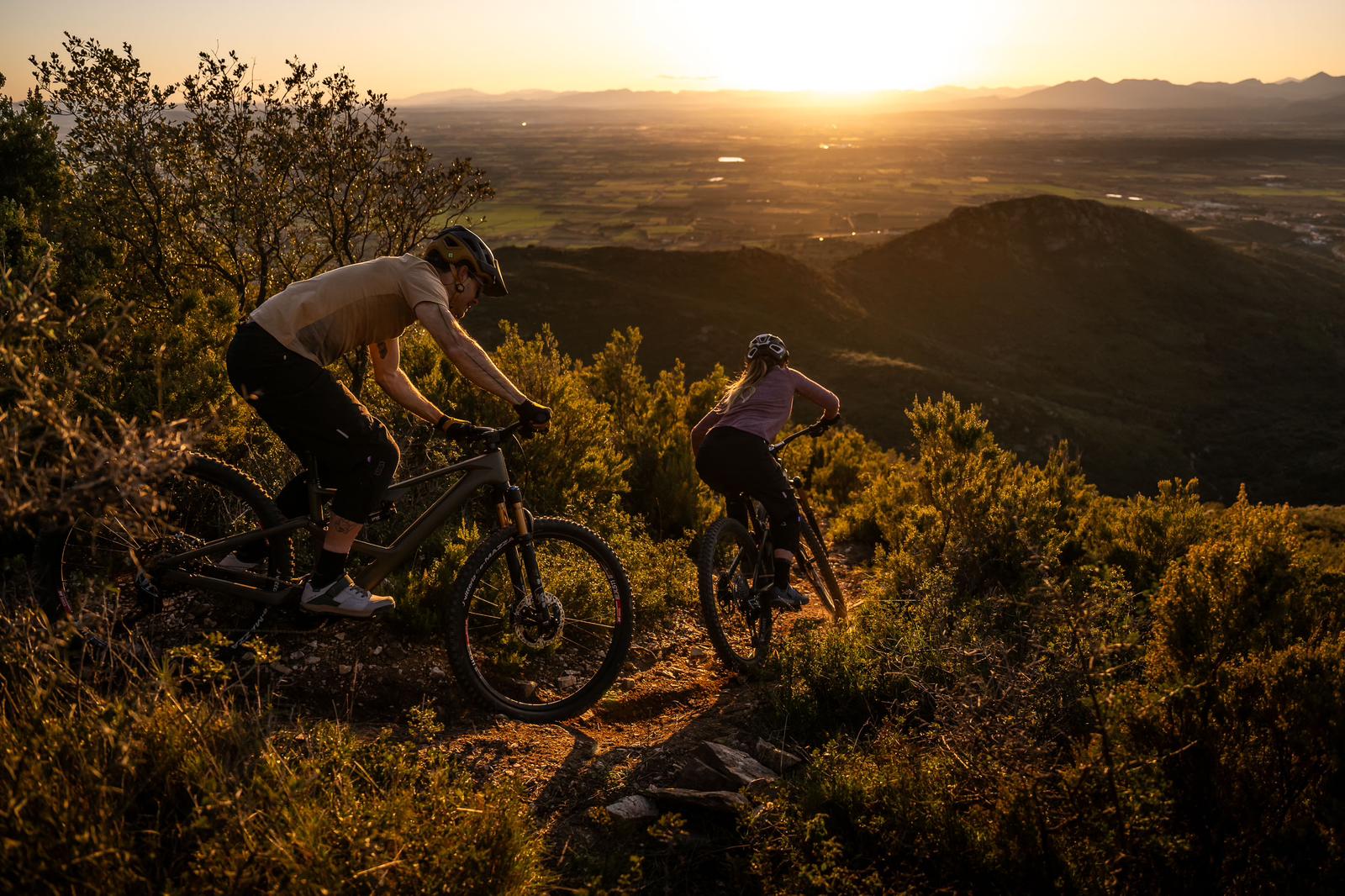 Mountain biking at sunset, two cyclists navigate a trail with expansive valley views, highlighting outdoor adventure and cycling in nature. Mountain Bike United.