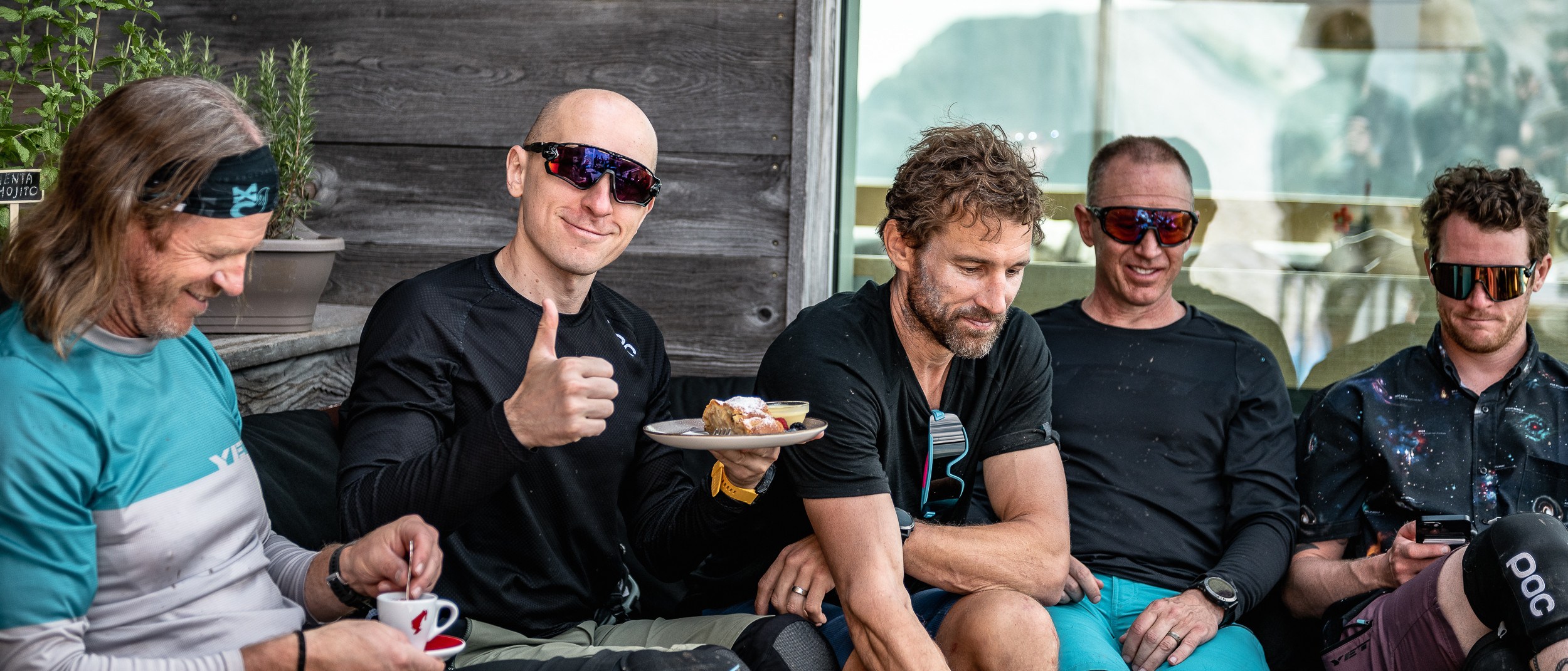Group of men in cycling gear enjoying a break with food and coffee, one giving a thumbs-up, wearing sunglasses, outdoor setting. Mountain Bike United.