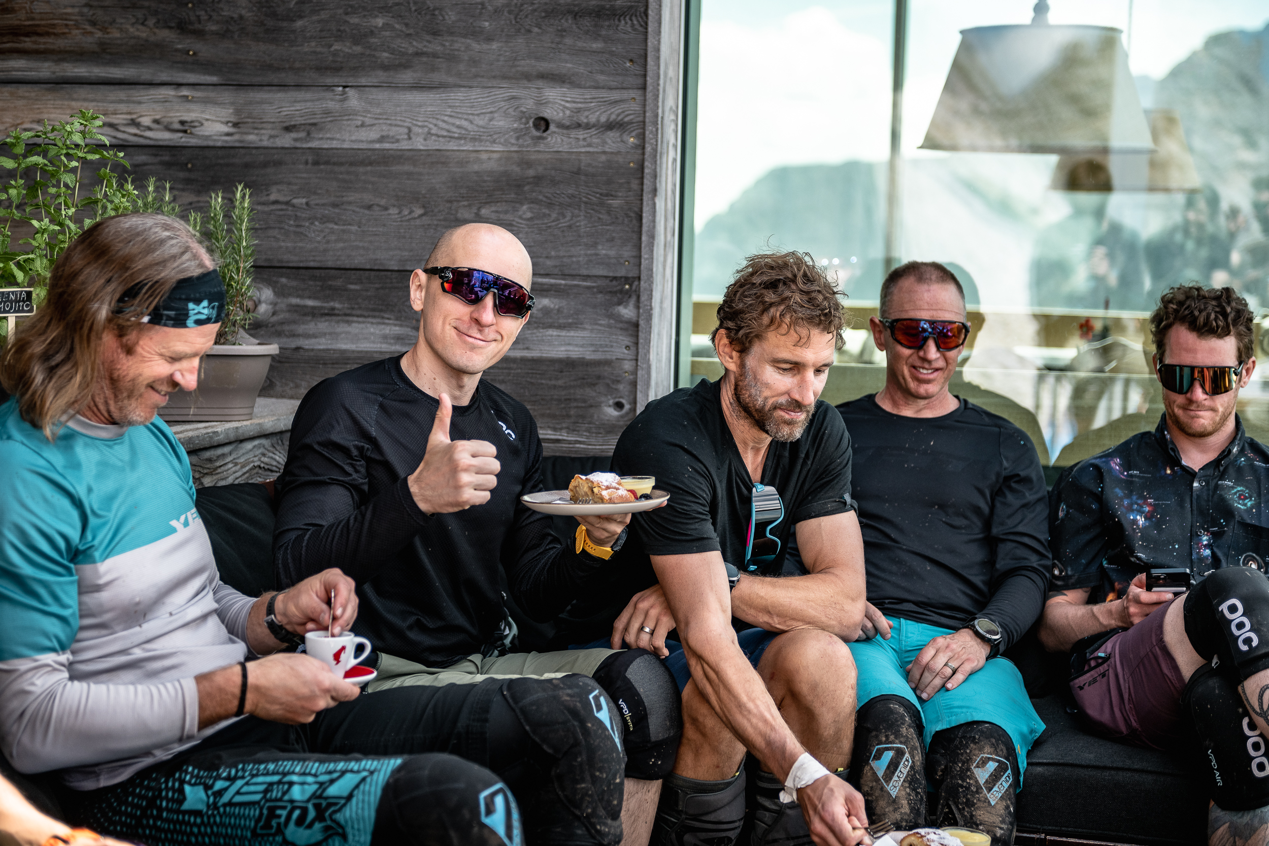 Group of men in cycling gear enjoying a break with food and coffee, one giving a thumbs-up, wearing sunglasses, outdoor setting. Mountain Bike United.
