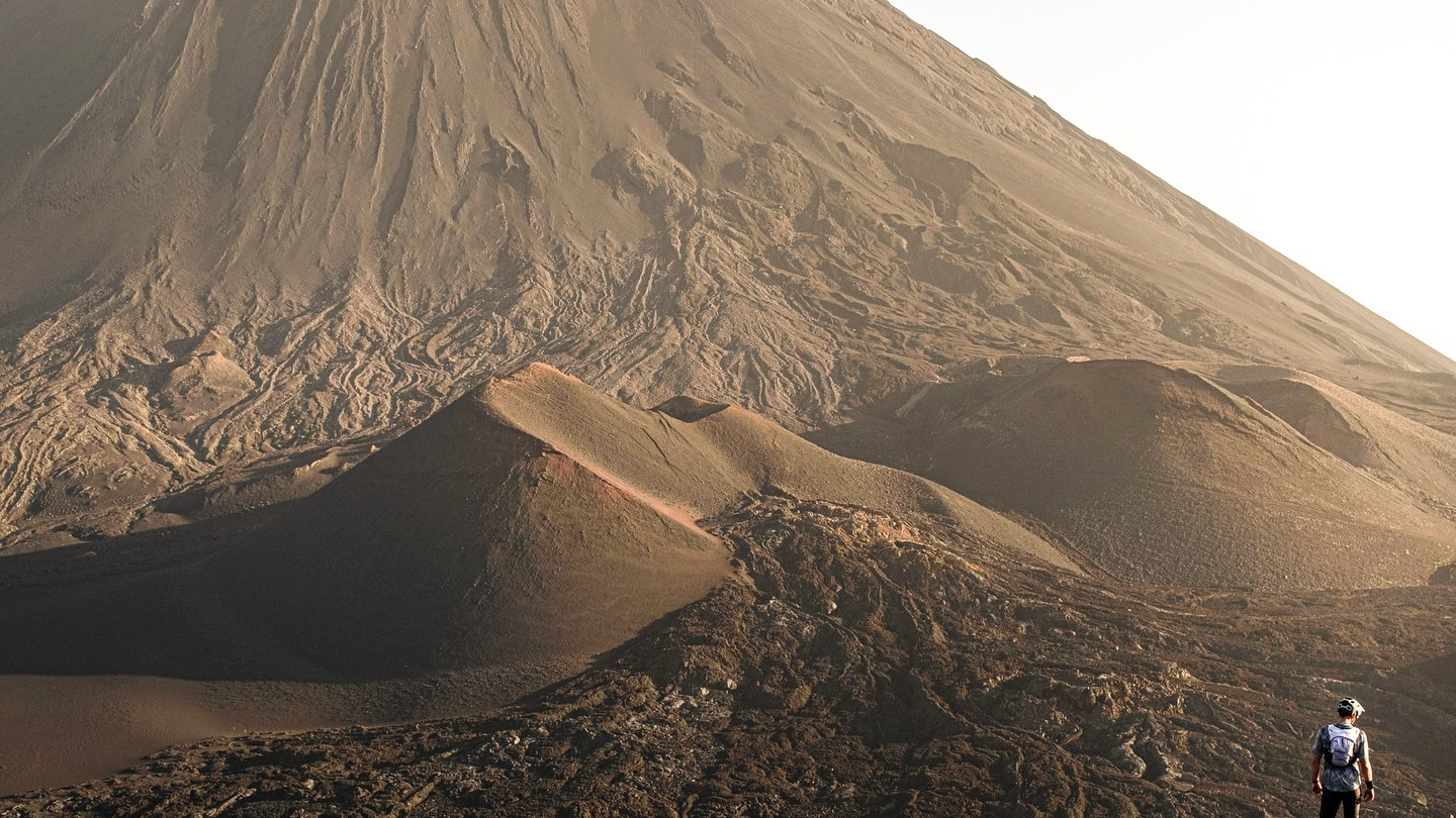 Hiker observing Mount Fogo's volcanic landscape in Cape Verde at sunrise, showcasing rugged terrain and natural beauty. Mountain Bike United.