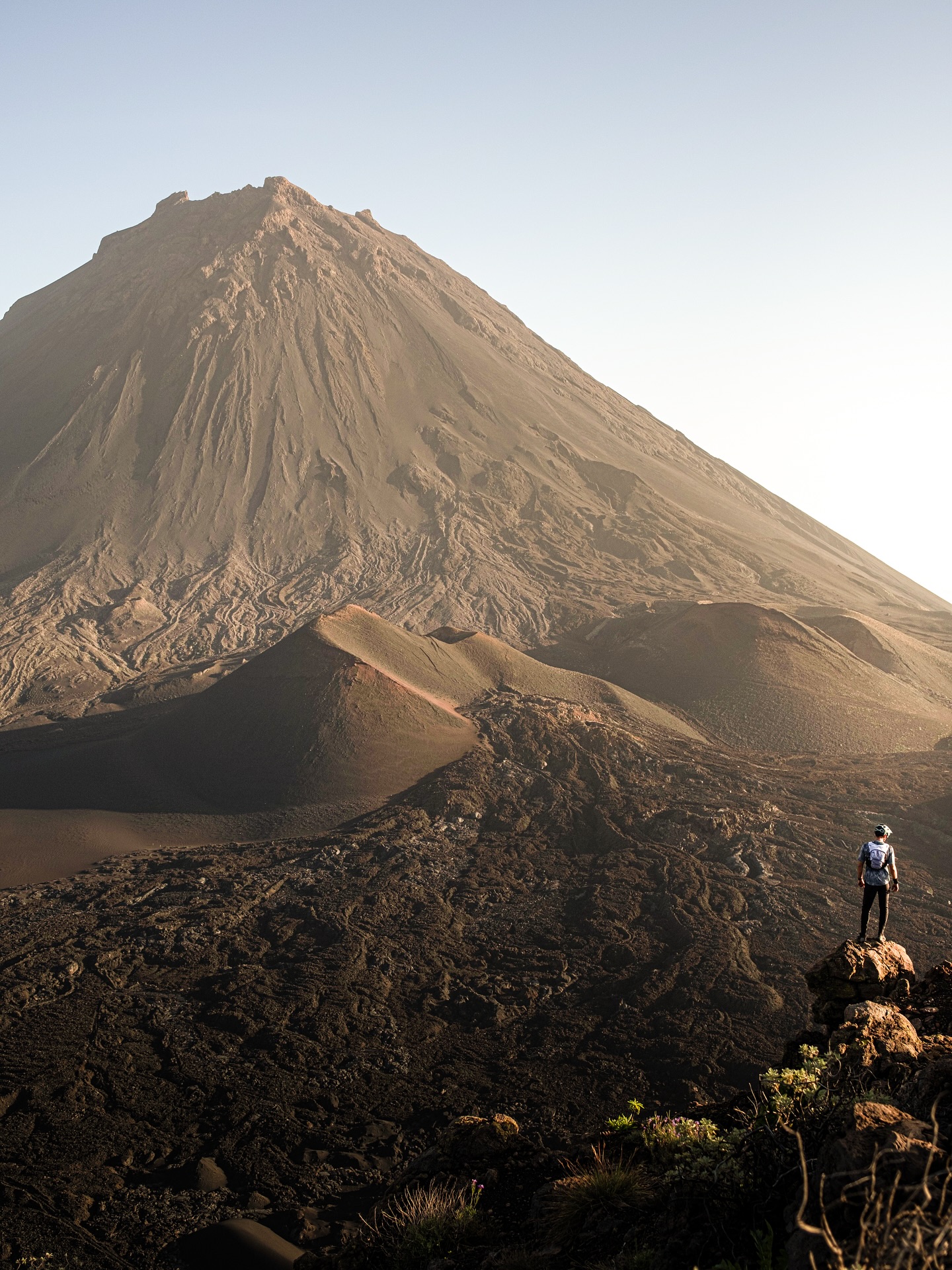 Hiker observing Mount Fogo's volcanic landscape in Cape Verde at sunrise, showcasing rugged terrain and natural beauty. Mountain Bike United.