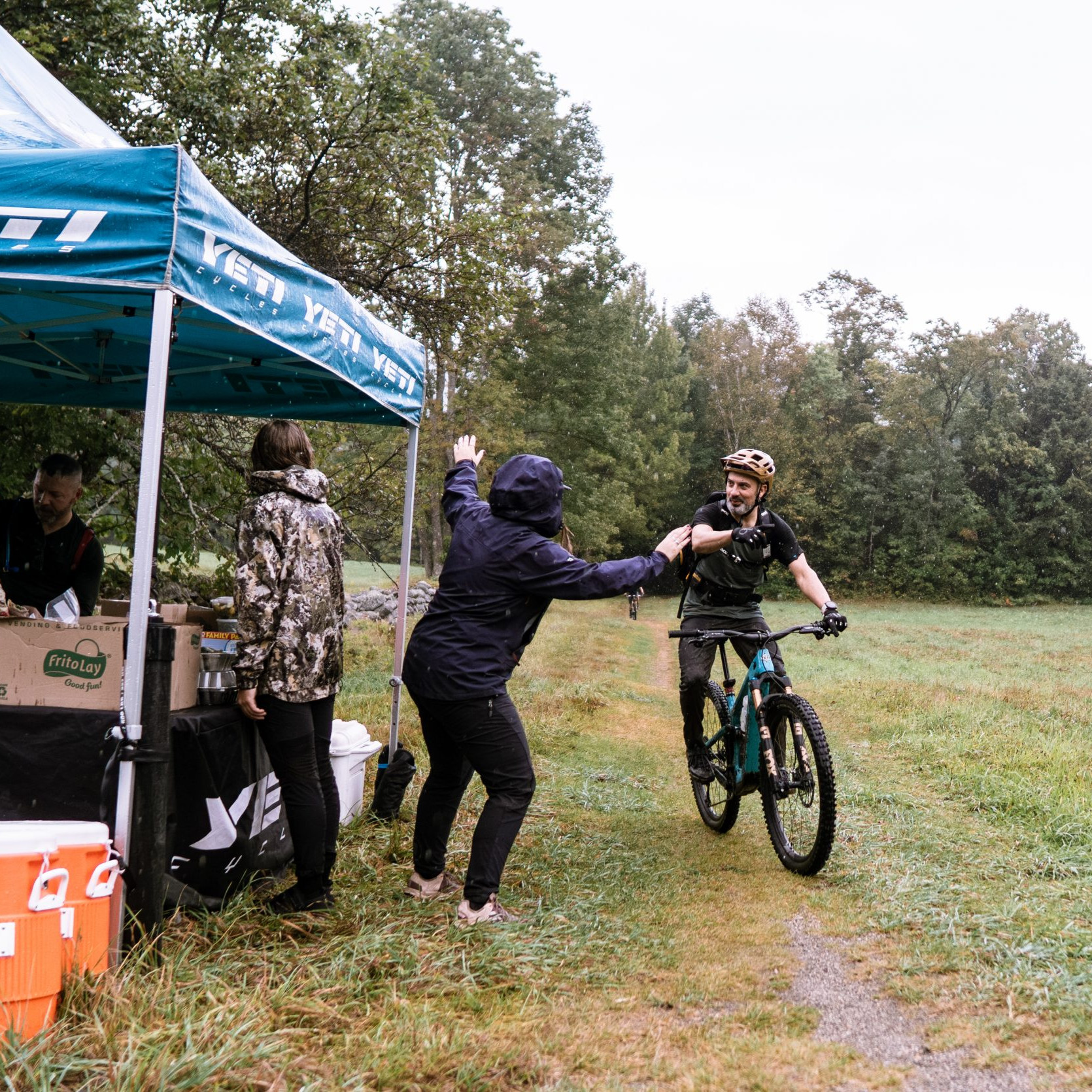 Mountain biker at MTB event stop, receiving aid from team under tent. Organise MTB event with on-site support for cyclists in scenic forest setting. Mountain Bike United.