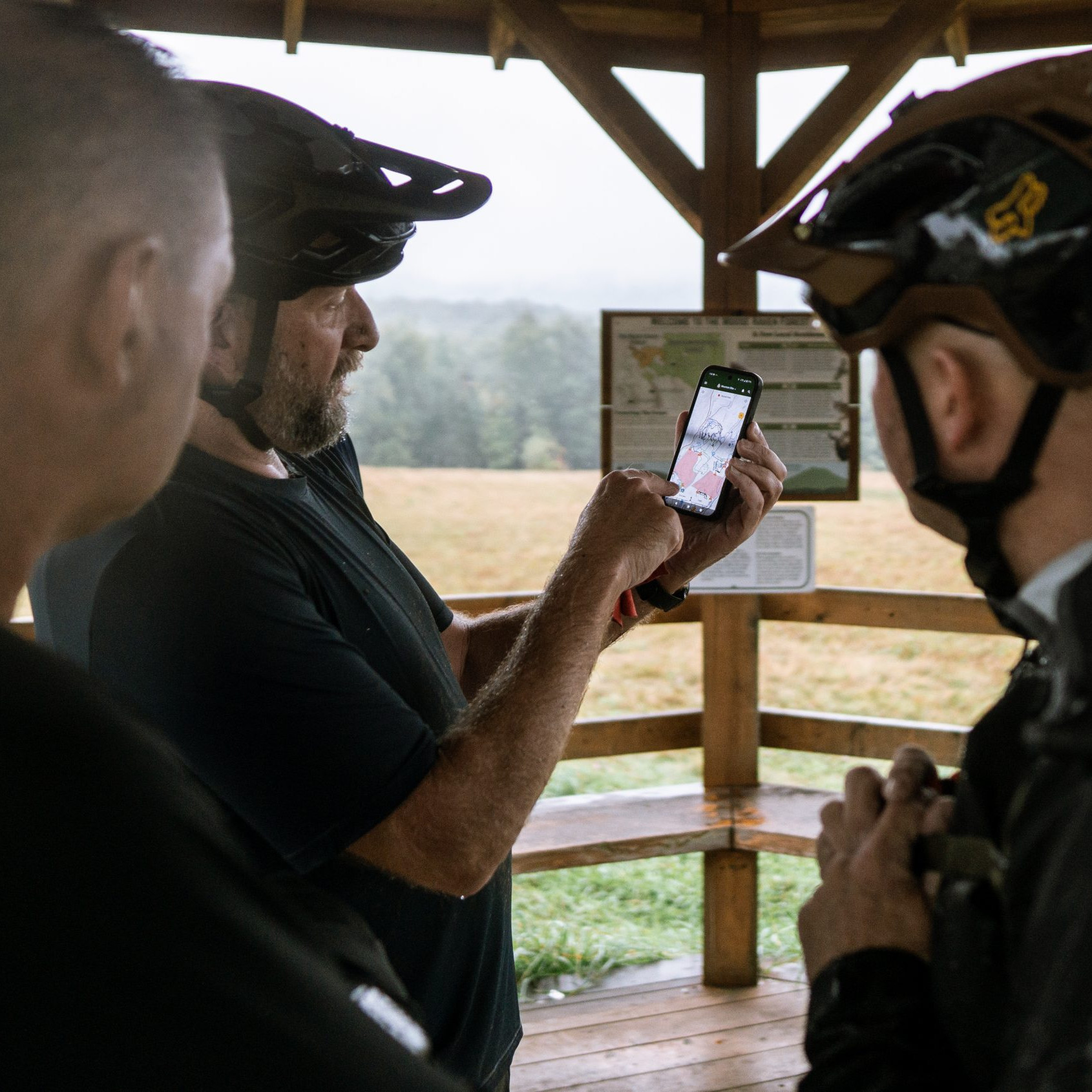 Cyclists in helmets planning to organise MTB event using a smartphone map under a wooden shelter in a scenic outdoor location. Mountain Bike United.