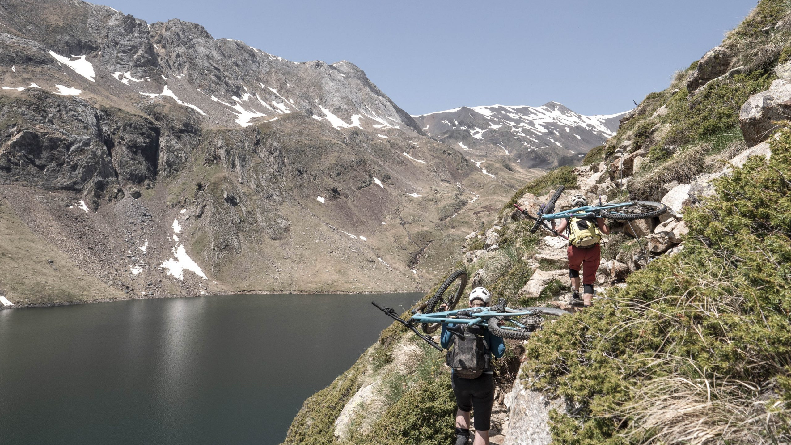 Hikers carrying bikes on a rugged trail in the Pyrenees with H+I Adventures, overlooking a mountain lake and peaks with patches of snow. Mountain Bike United.