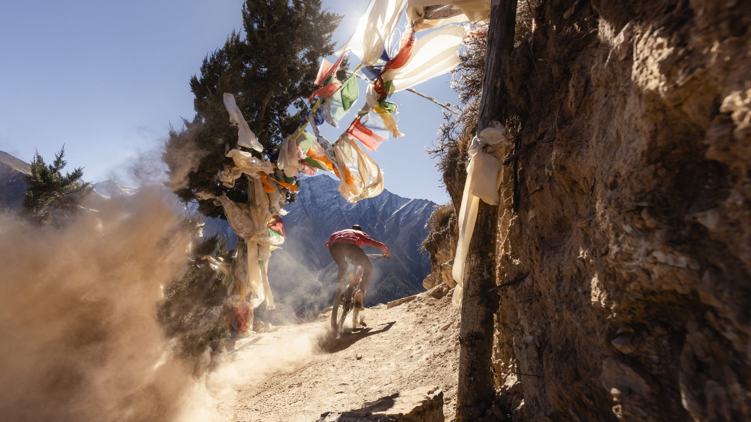 Mountain biker rides downhill trail with dust cloud and prayer flags, featuring Kilian Bron's Mandala performance. Perfect for adventure enthusiasts. Mountain Bike United.