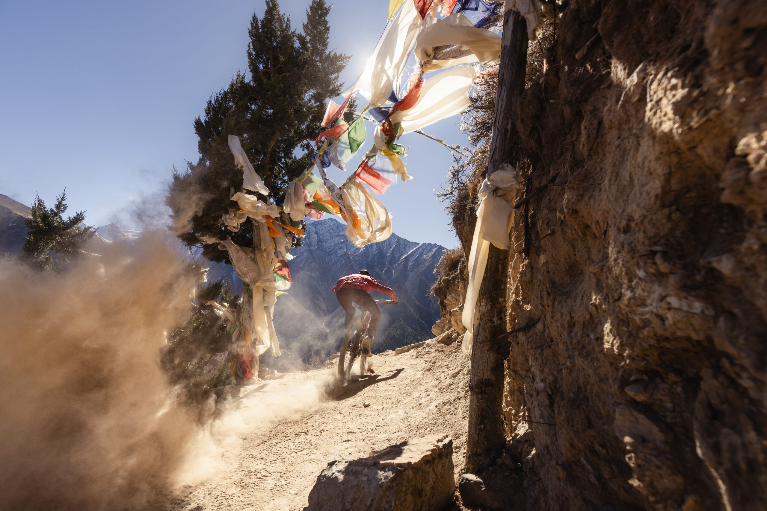 Mountain biker rides downhill trail with dust cloud and prayer flags, featuring Kilian Bron's Mandala performance. Perfect for adventure enthusiasts. Mountain Bike United.