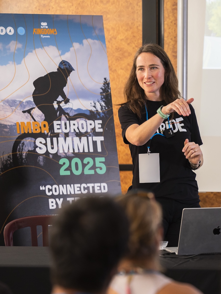 Speaker presenting at IMBA Europe Summit 2025 with event poster in background. Mountain Bike United.